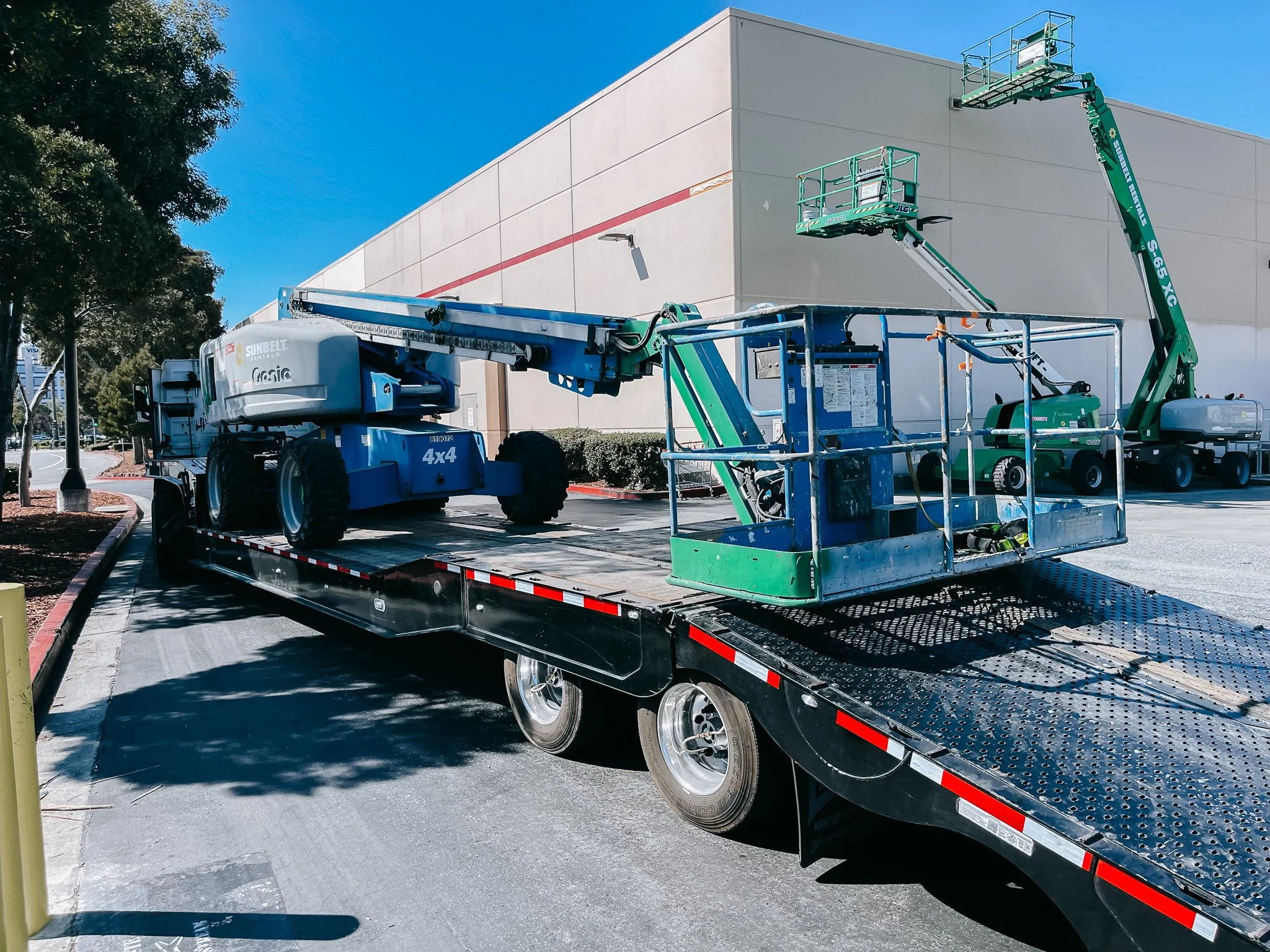 A flatbed truck carrying a green telescopic boom lift with two work platforms, parked on a street near a modern building and trees.
