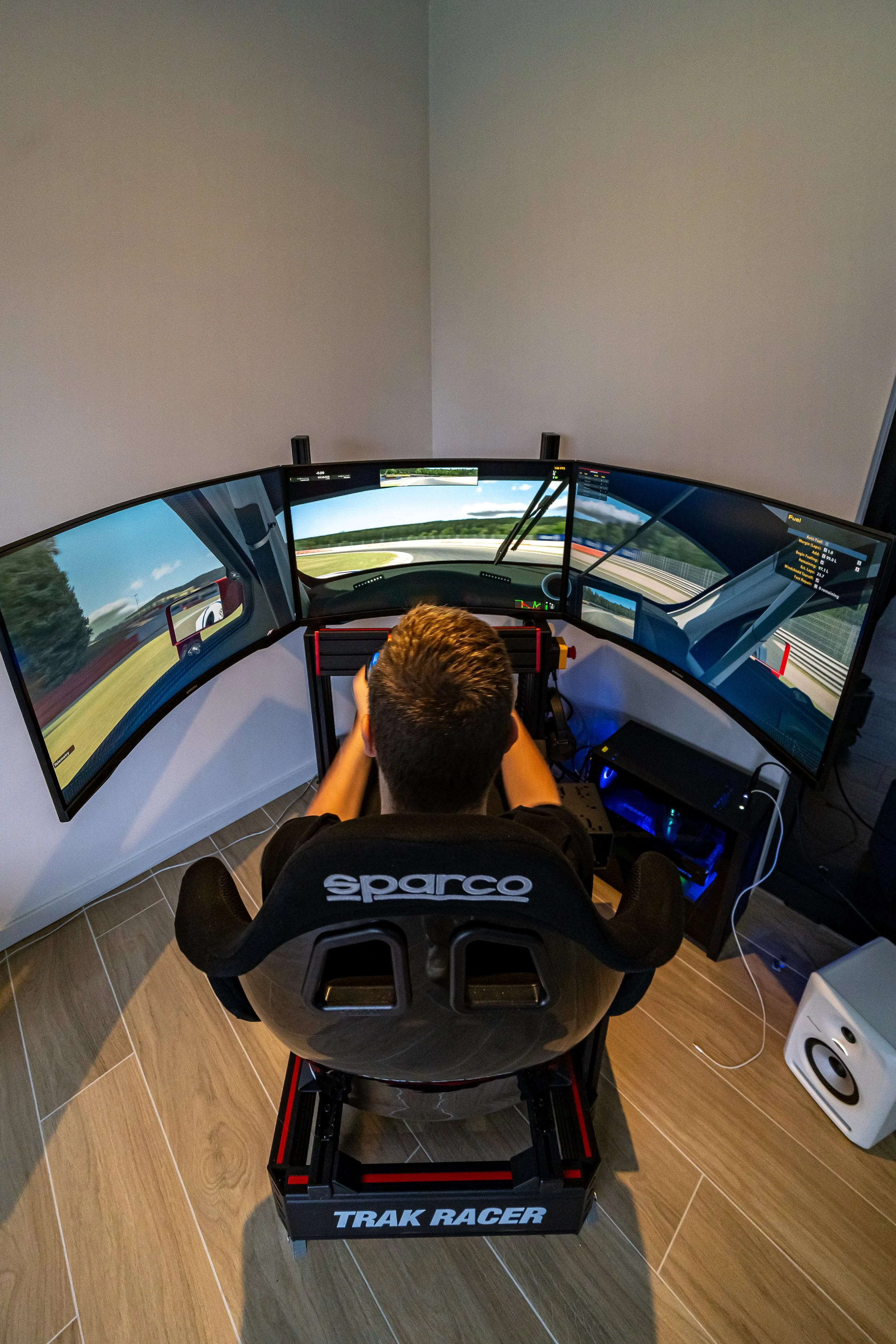 Persoon die achter een racestuur zit, omgeven door drie computerschermen die een racegame op een racetrack weergeven