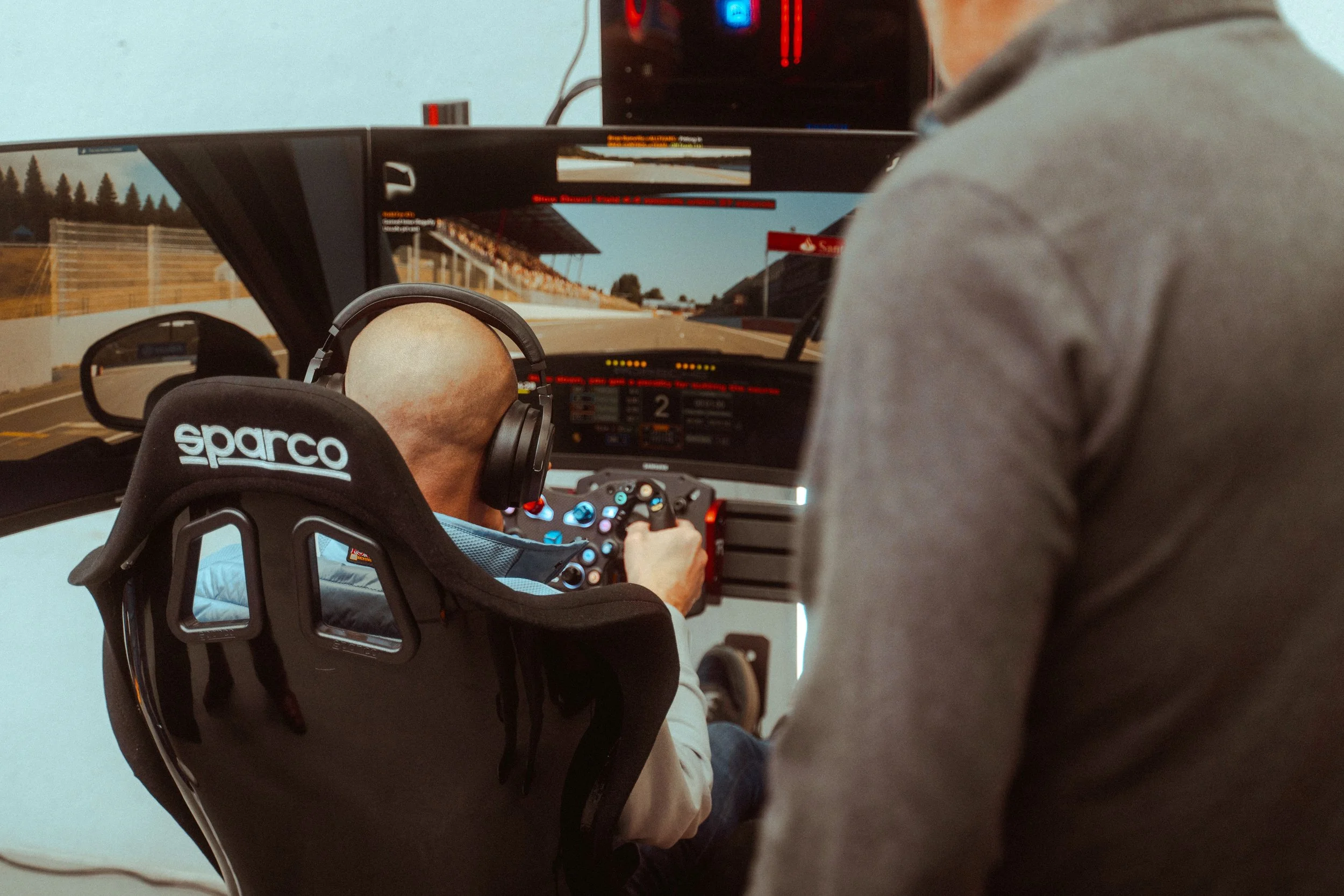 Man zit in een race-autotechniek simulator met meerdere schermen en koptelefoon, terwijl iemand anders naast hem staat en het stuur vasthoudt.