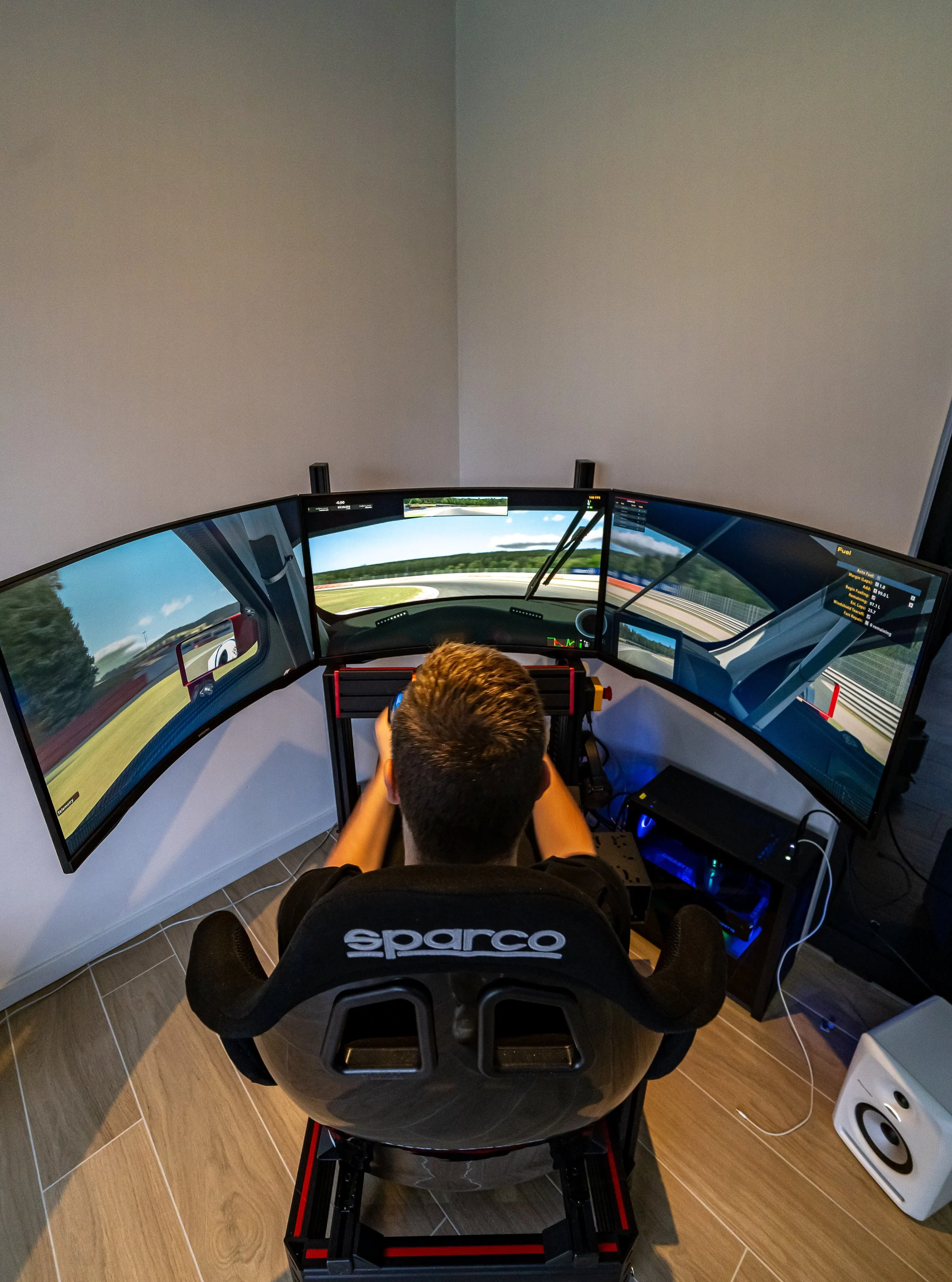Een persoon zit in een race simulator met drie schermen die een racebaan op een computerscherm tonen, in een kamer met houten vloeren en een witte muur.