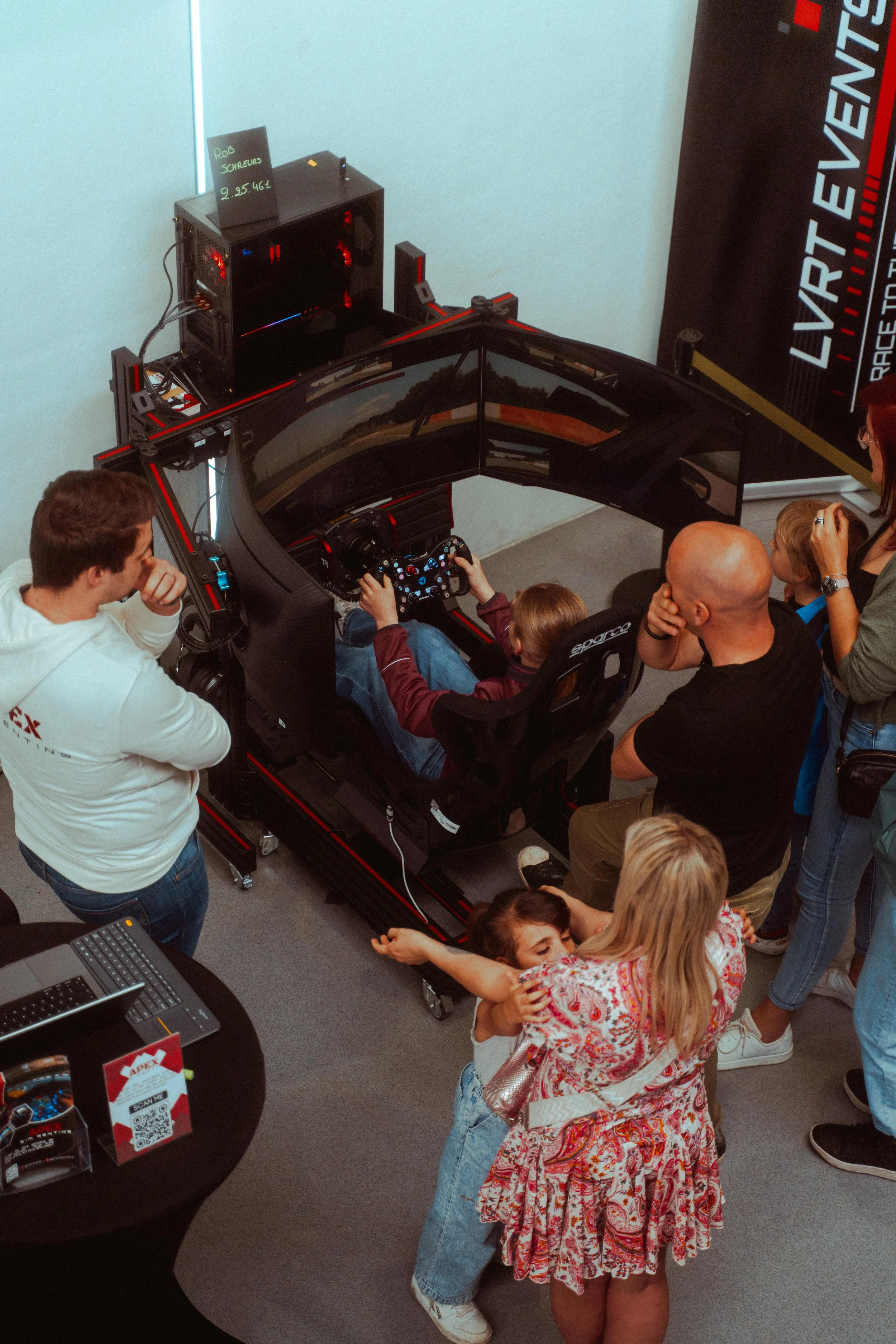 Mensen kijken naar een racegame simulator met een kind dat aan het sturen is, en een vrouw die het kind omhelst.