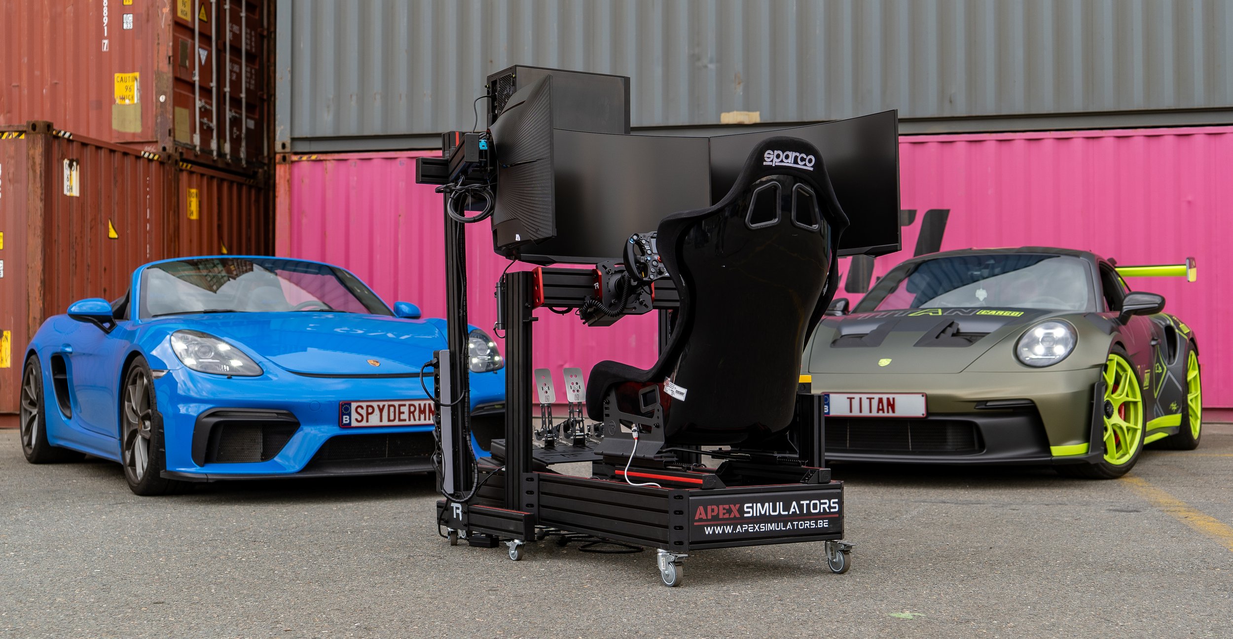 Twee sportwagens, een blauwe Porsche en een grijs-groene Porsche, staan op een parkeerplaats voor een rijcontainer. Tussen de auto's staat een race-simulatorset met een zwart racestoeltje en drie grote schermen.