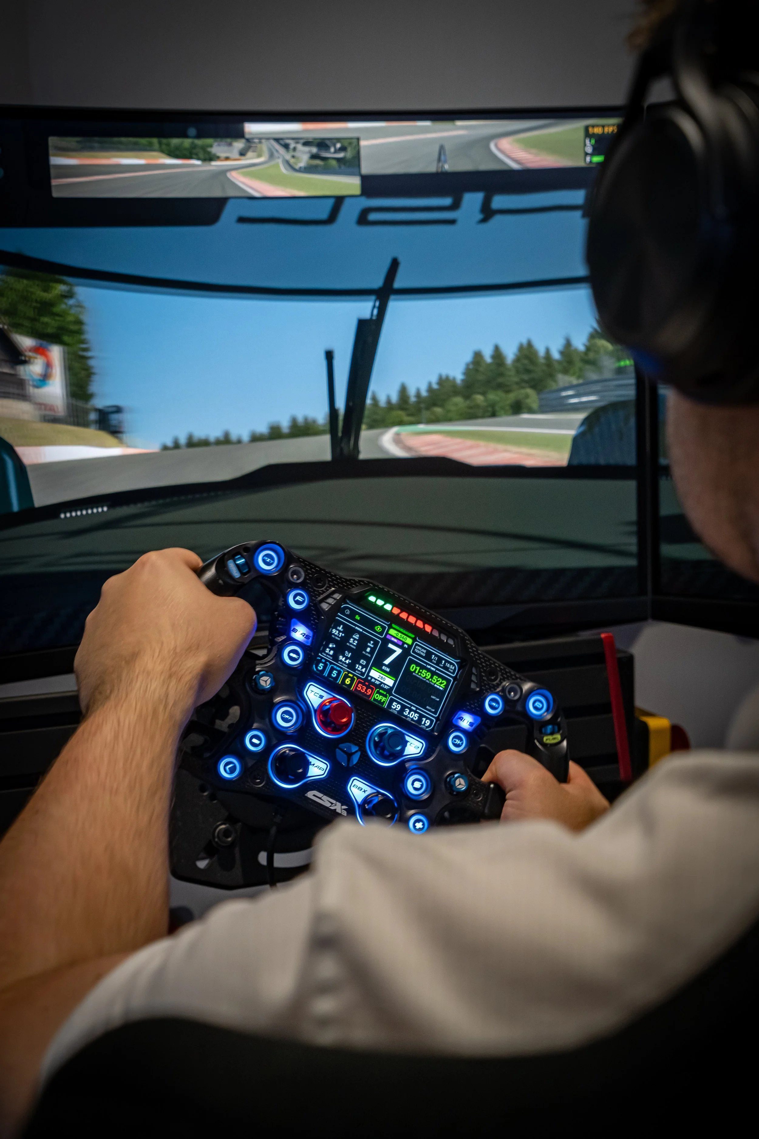 Een coureur omgeven door een simulatie met een raceauto, met een stuurwiel en digitale schermen, onderweg op een racetrack met bochtige weg en groene bomen.