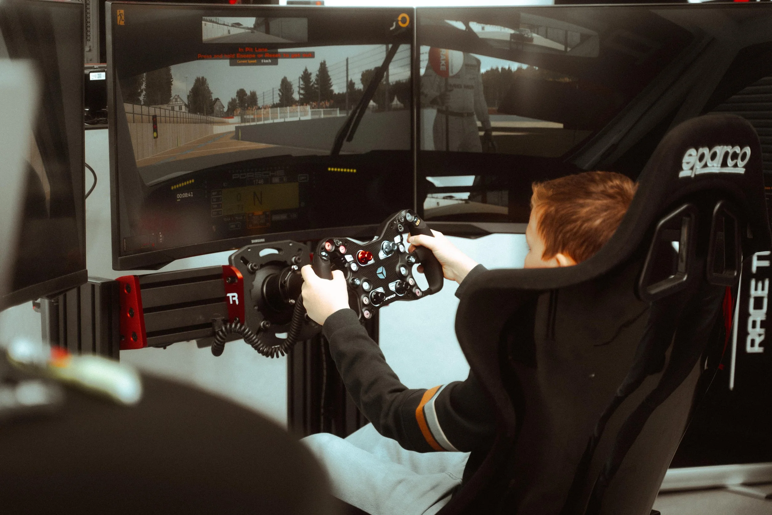 Een kind zit in een raceauto simulatorsetup, met een racestuur en meerdere schermen die een racegame tonen.