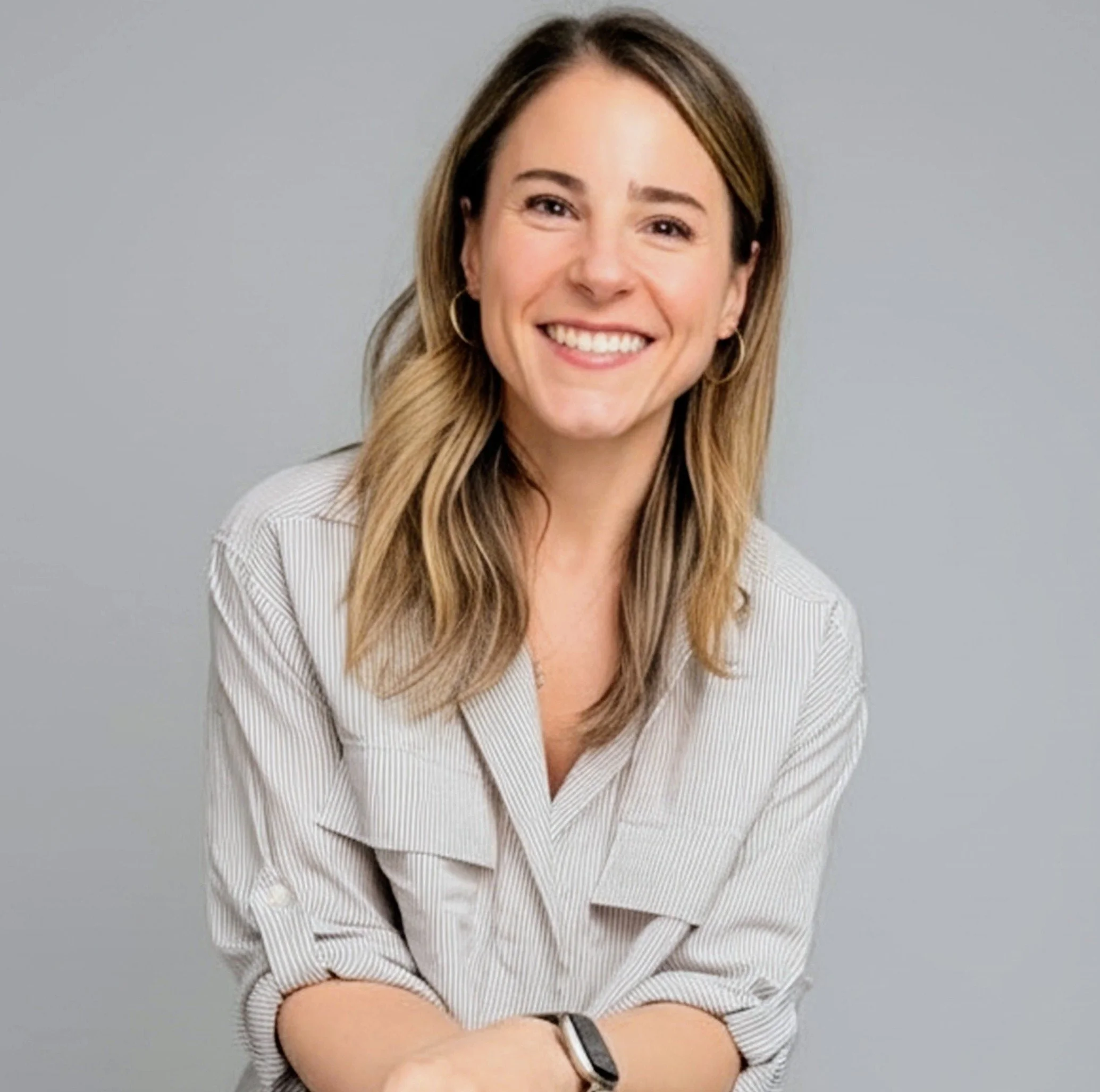 A smiling woman with long, wavy blonde hair, wearing a light-colored, striped dress shirt and gold hoop earrings, crossing her arms against a plain grey background.