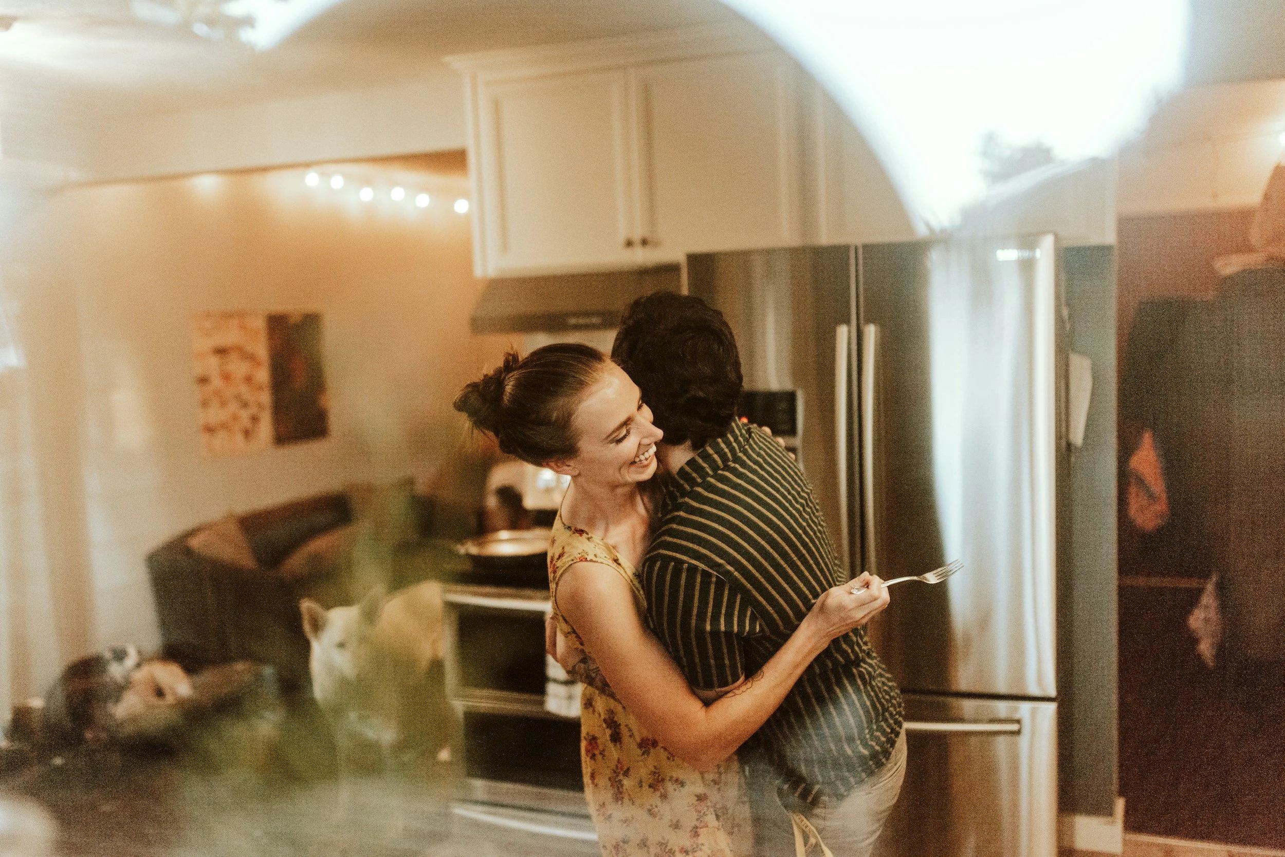 A woman smiling and hugging a man in a kitchen, holding a fork. There are dogs on the counter in the foreground, and a fridge and cabinets in the background.