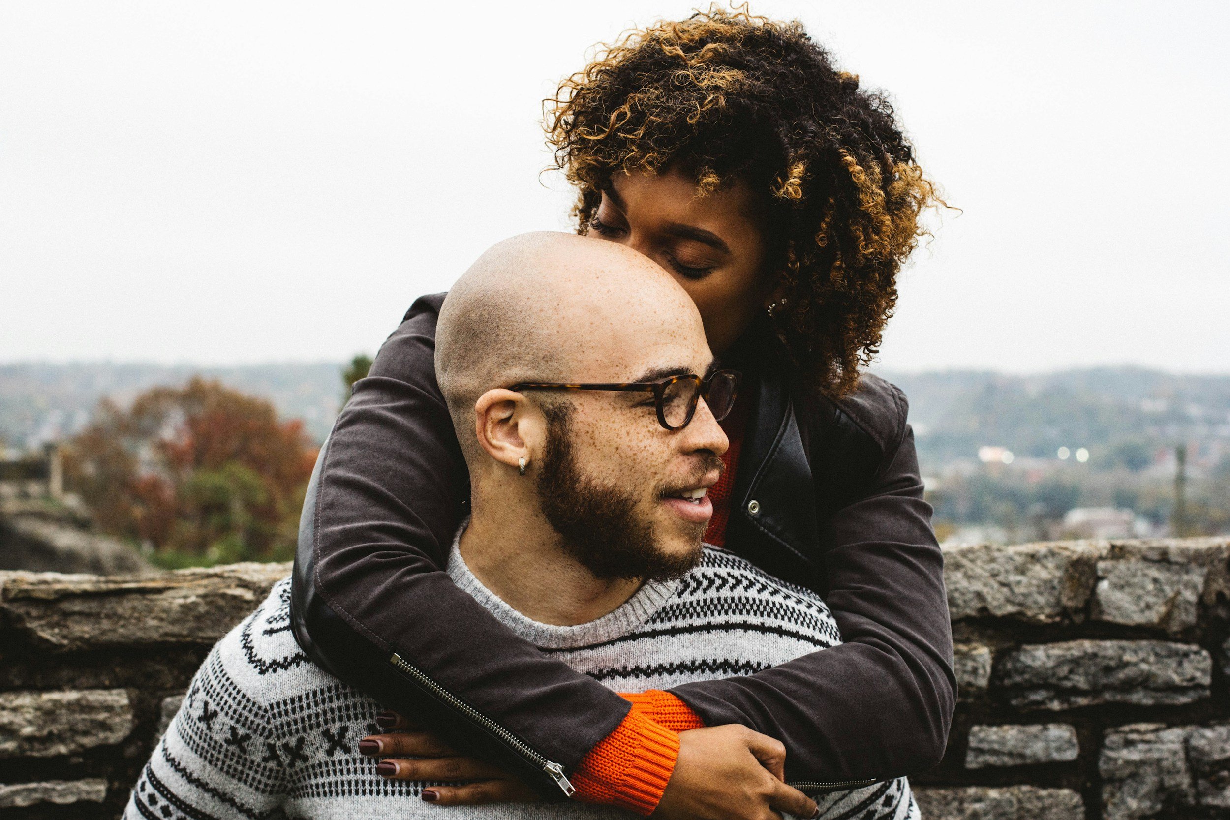 A woman with curly hair hugging a bald man with glasses, both smiling and enjoying each other's company outdoors with a blurred background of trees and hills.