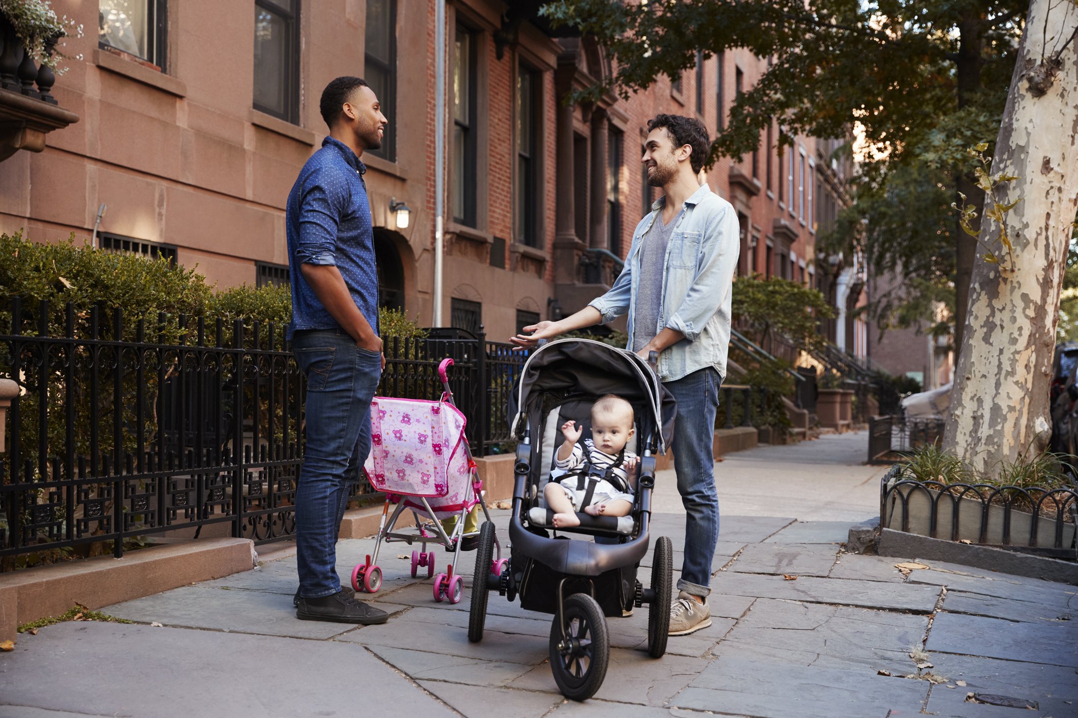 Two men chatting on a city sidewalk beside a baby in a stroller and a pink toy stroller.