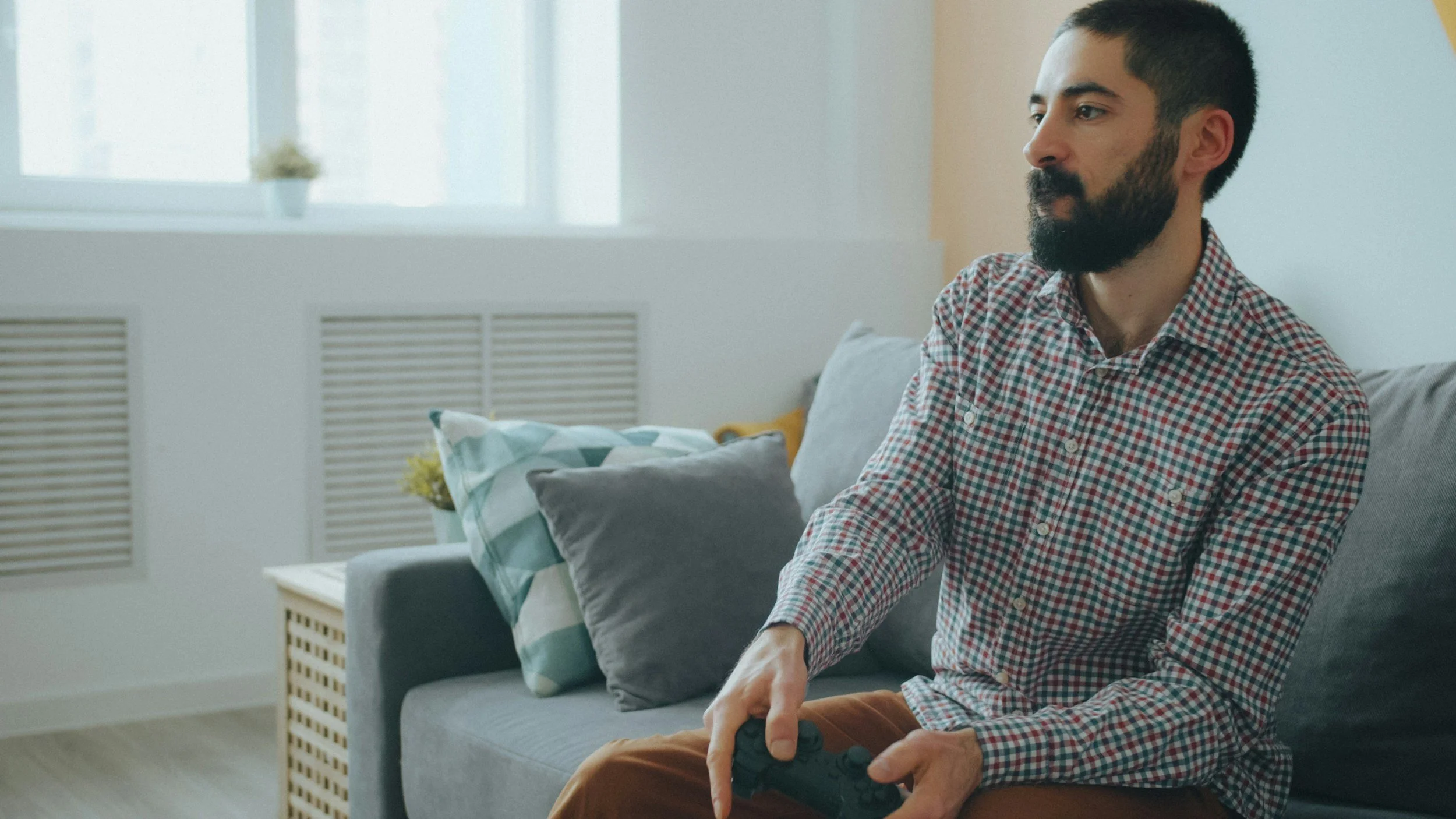 Bearded man in a plaid shirt holding a video game controller while sitting on a couch at home