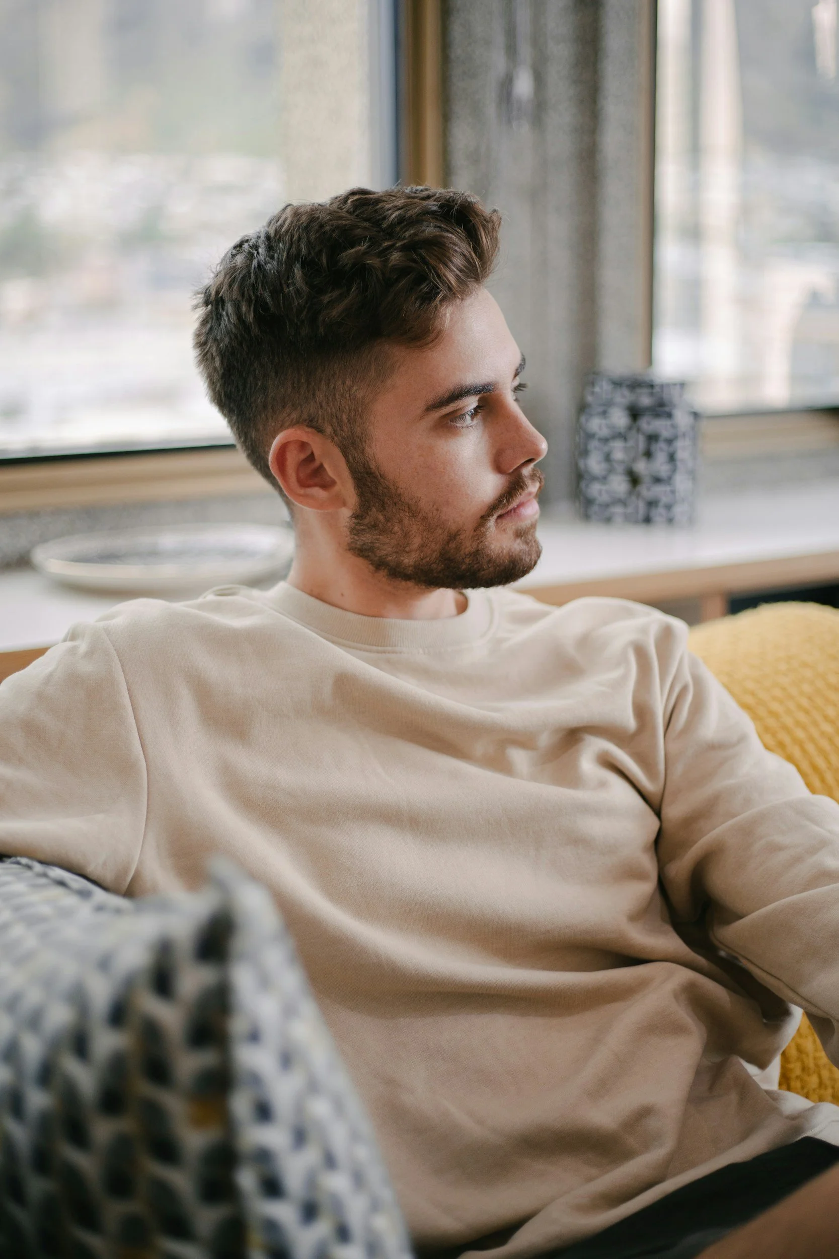 Bearded man in a beige sweatshirt relaxing on a couch beside a window