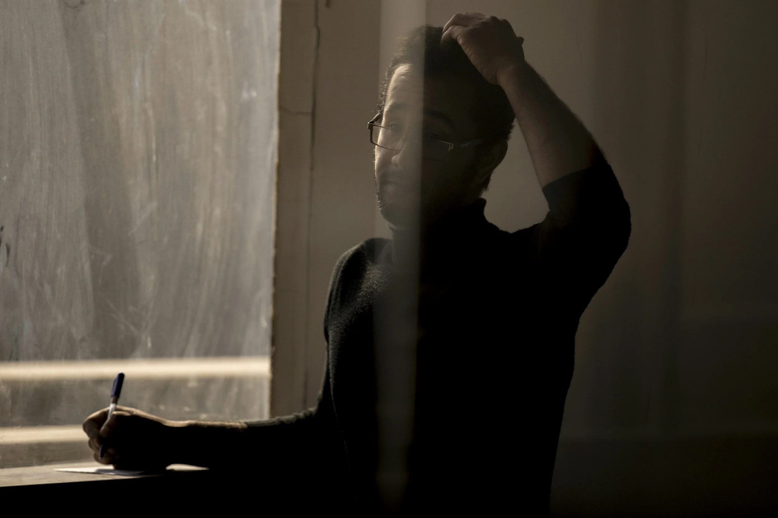 Man wearing glasses rubs his head while holding a pen and notebook in dim light.