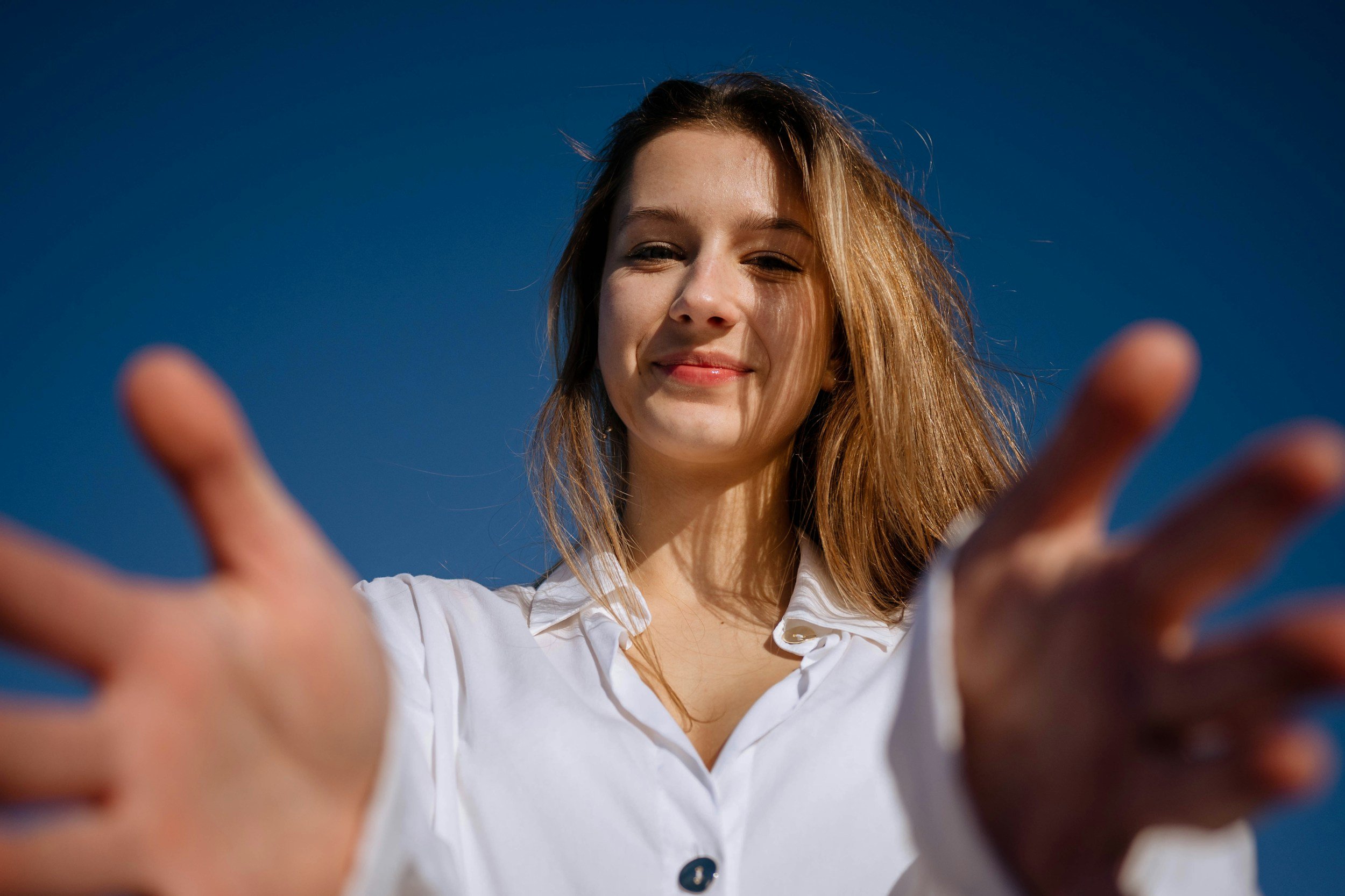 Smiling woman in a white blouse reaching toward the camera against a blue sky