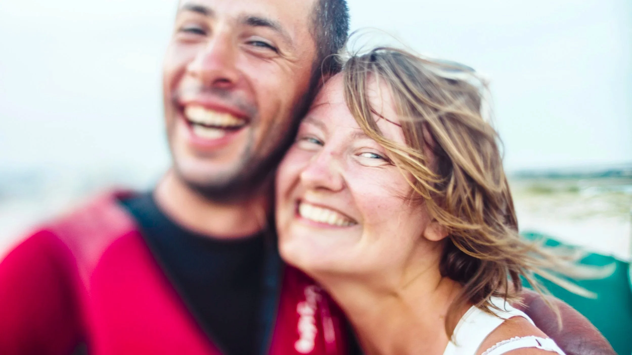 Close-up of a smiling man and woman hugging outdoors, with the ocean in the background.