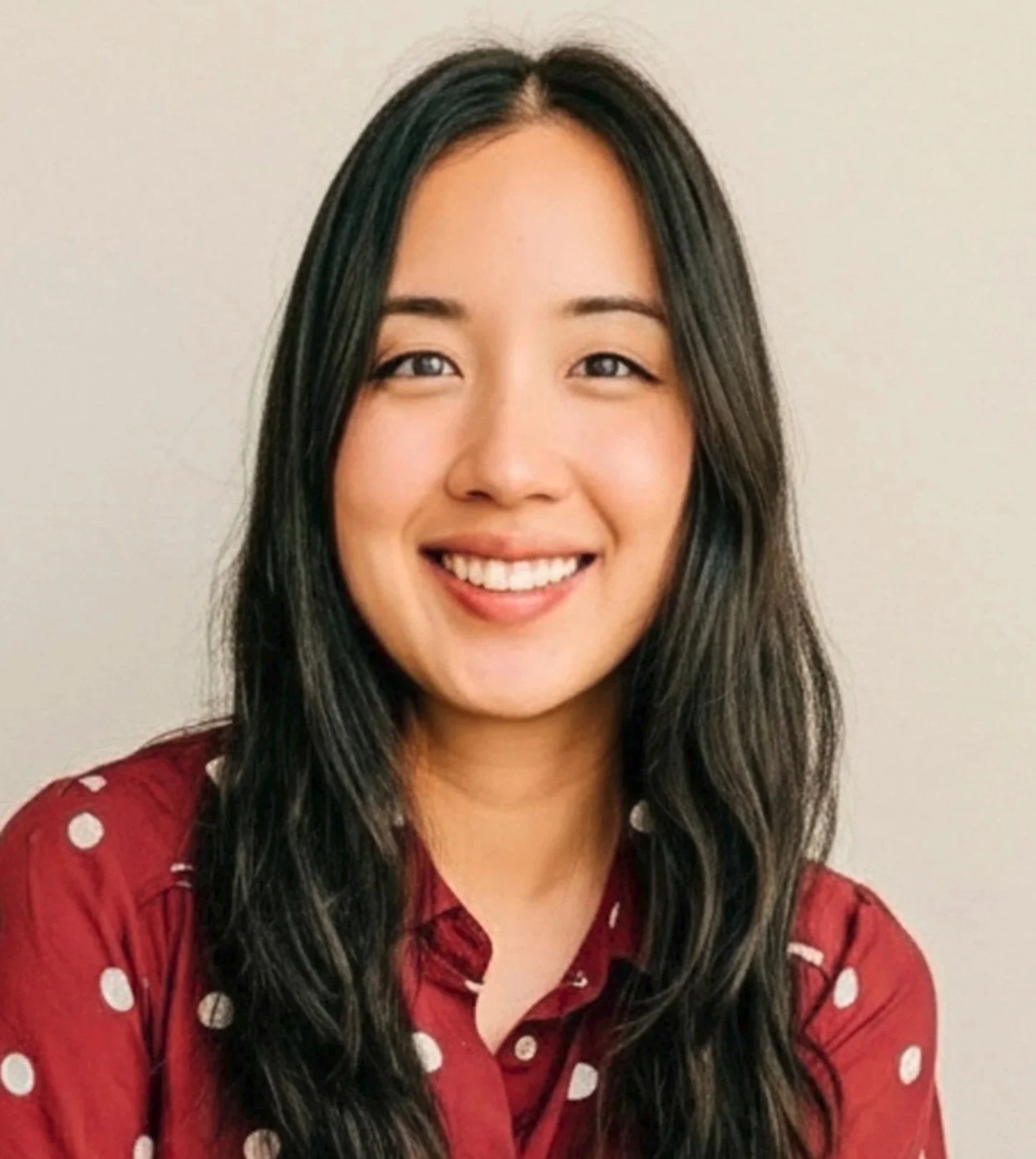 A close-up portrait of a smiling young woman with long black hair, wearing a black and gray patterned top, against a plain light gray background.