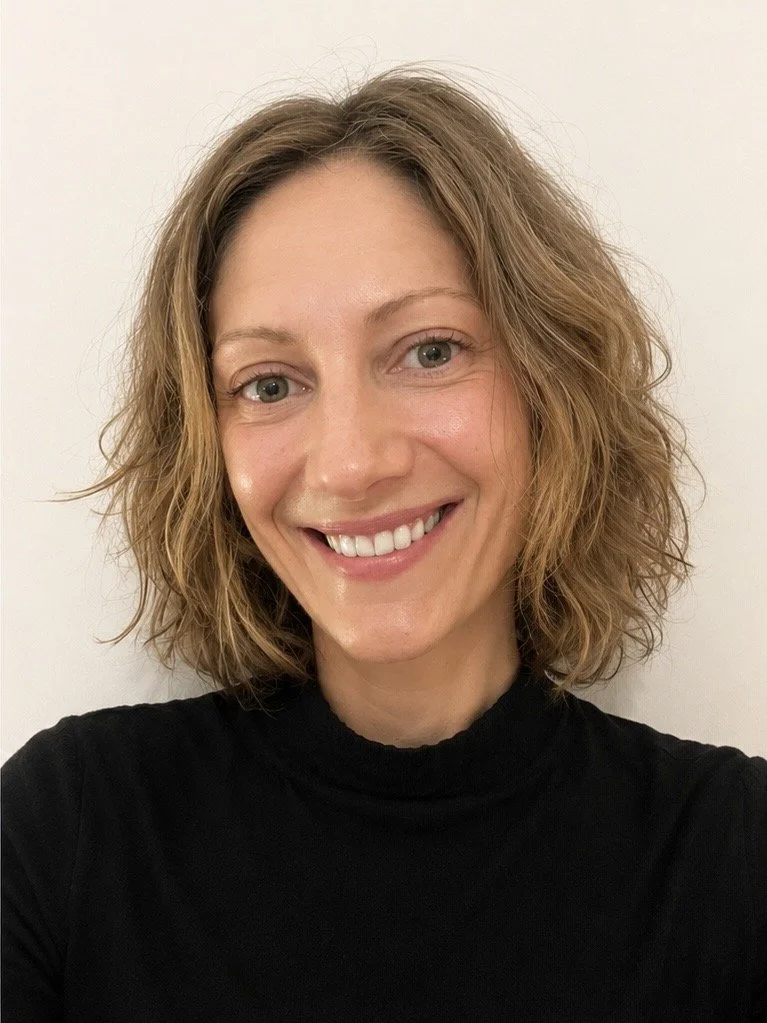 A woman with curly, shoulder-length hair smiling at the camera, wearing a black top against a plain light-colored background.