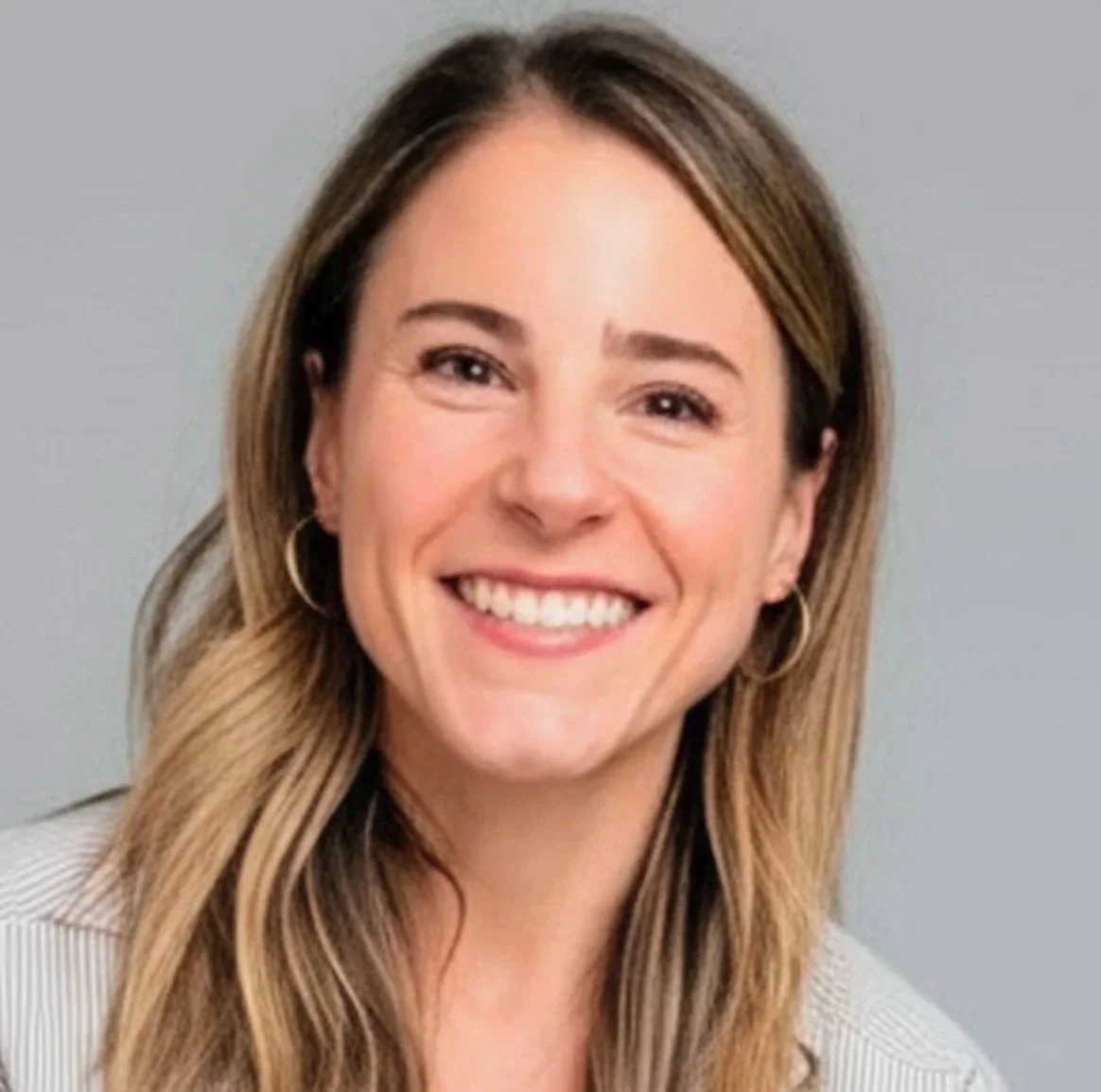 Close-up portrait of a smiling young woman with shoulder-length wavy brown hair, wearing a black top and a pendant necklace, against a plain white background.