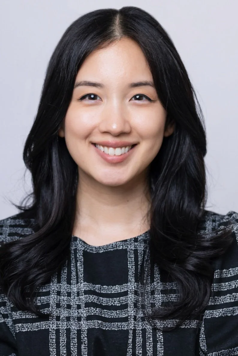 A young woman with long black hair, smiling and wearing a black and gray patterned top.