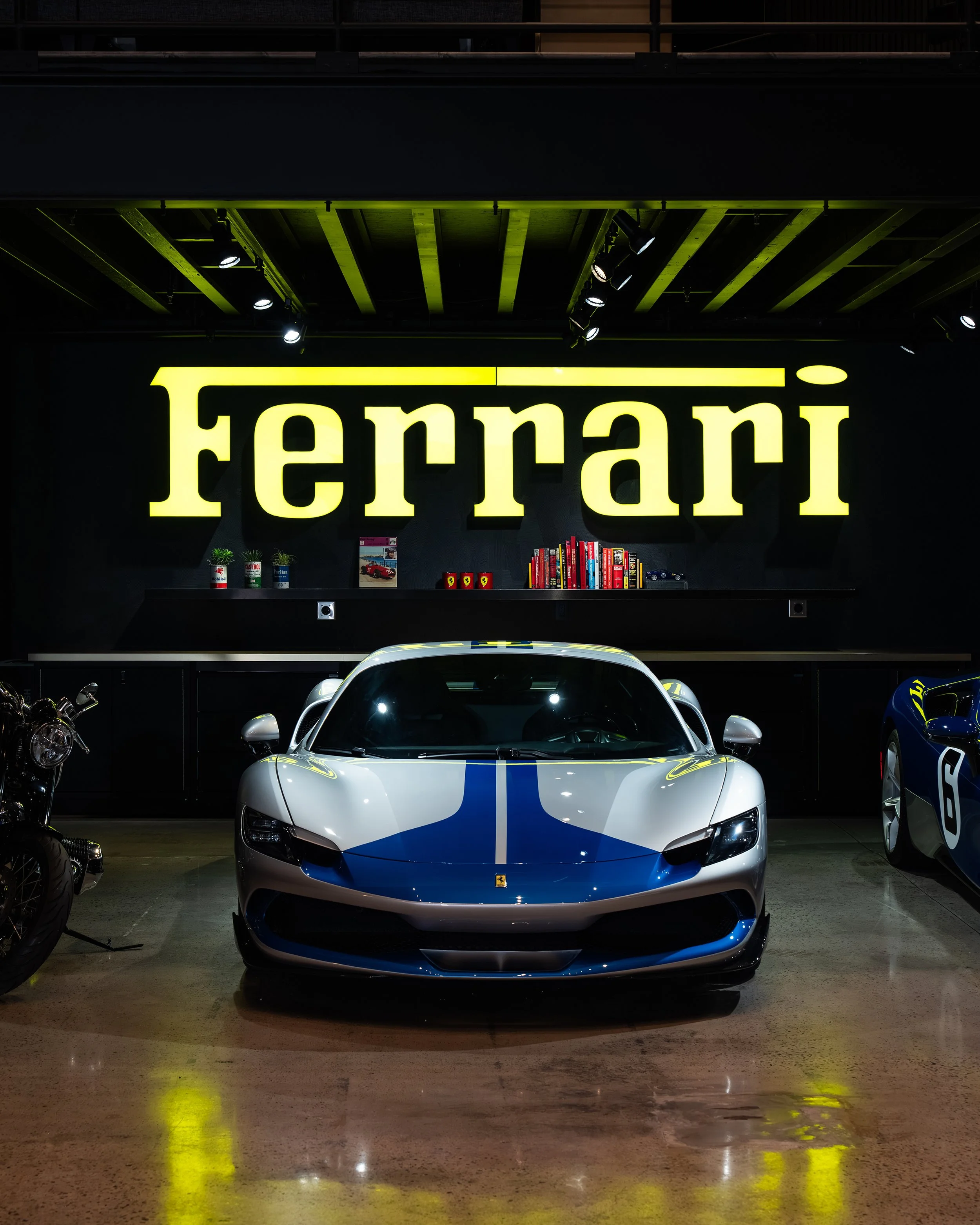 A white and blue Ferrari sports car is displayed in front of a large illuminated yellow Ferrari sign inside a showroom. The background has a black wall with a shelf holding small plants, books, and memorabilia.