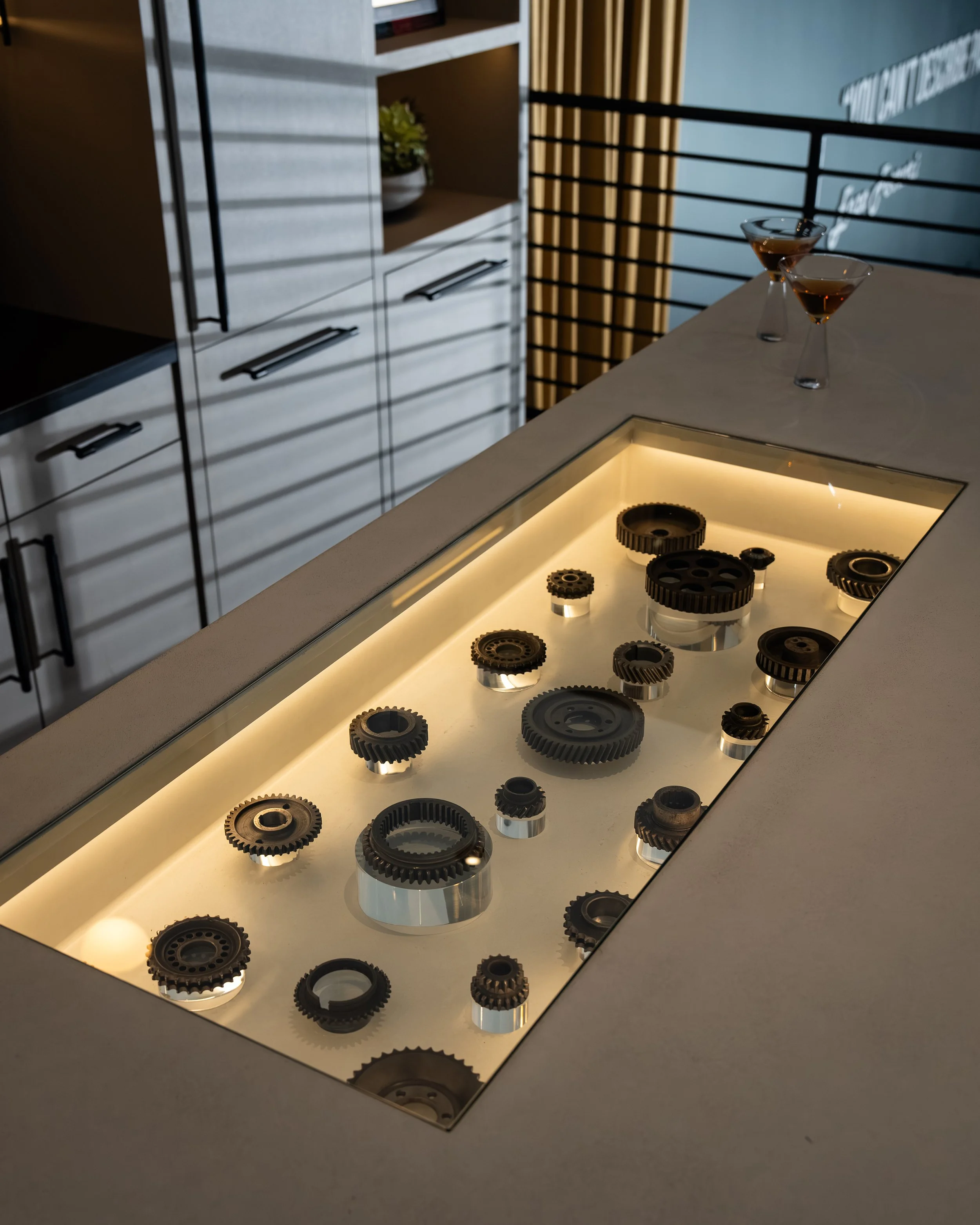 Display of various metal gears under glass on a modern table in a stylish, dimly lit room with two cocktail glasses in the background.