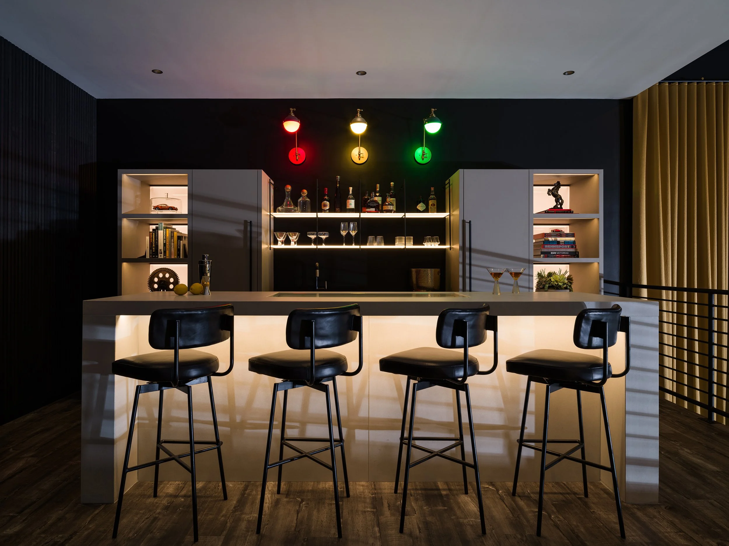 Modern home bar with black, white, and gold accents, featuring four black leather bar stools, a white illuminated island, and a black wall with colorful lighting and shelves with books and decor.  Traffic light red, yellow, green illumination effect.