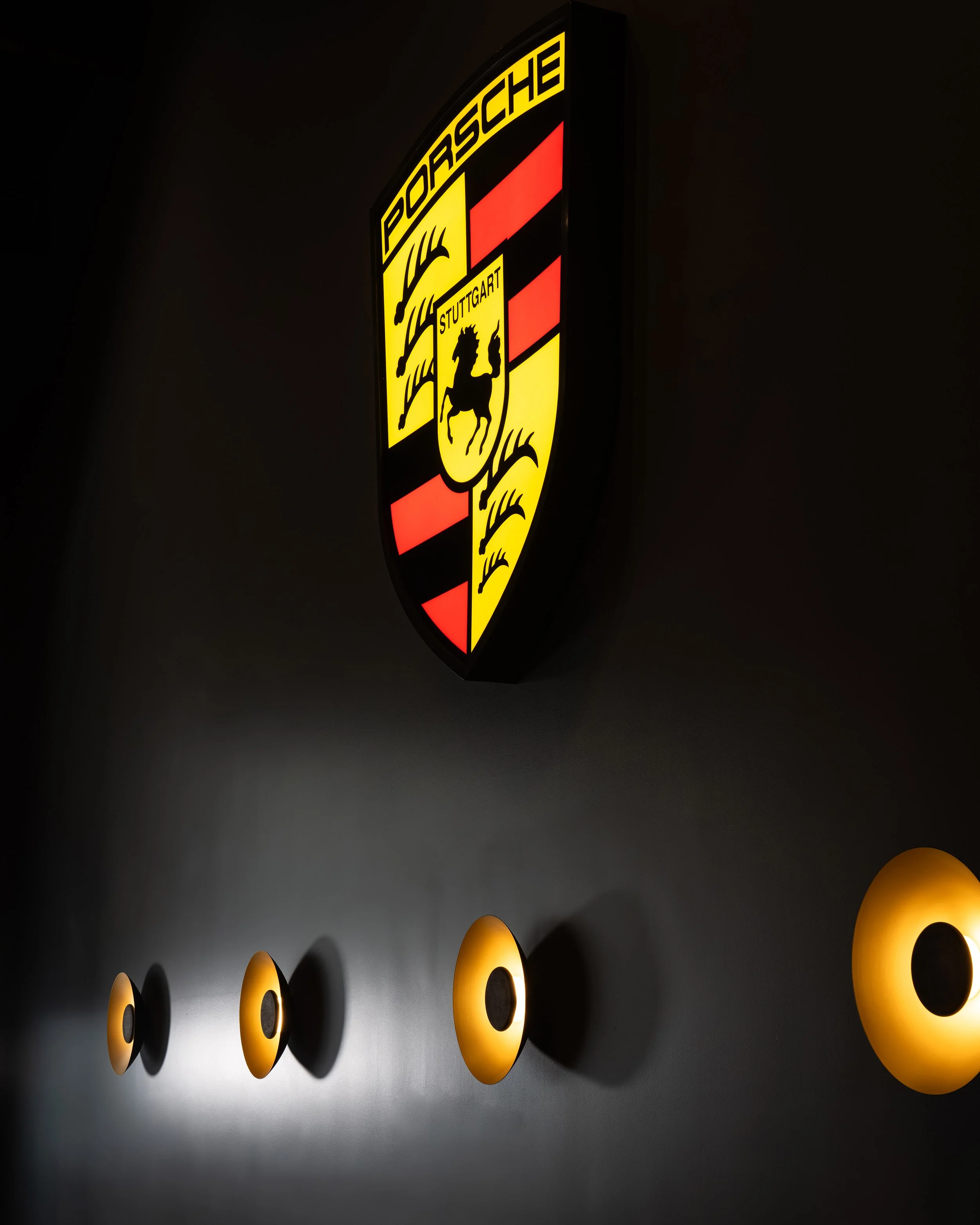 Porsche emblem illuminated on a dark wall with four circular yellow wall lights below.
