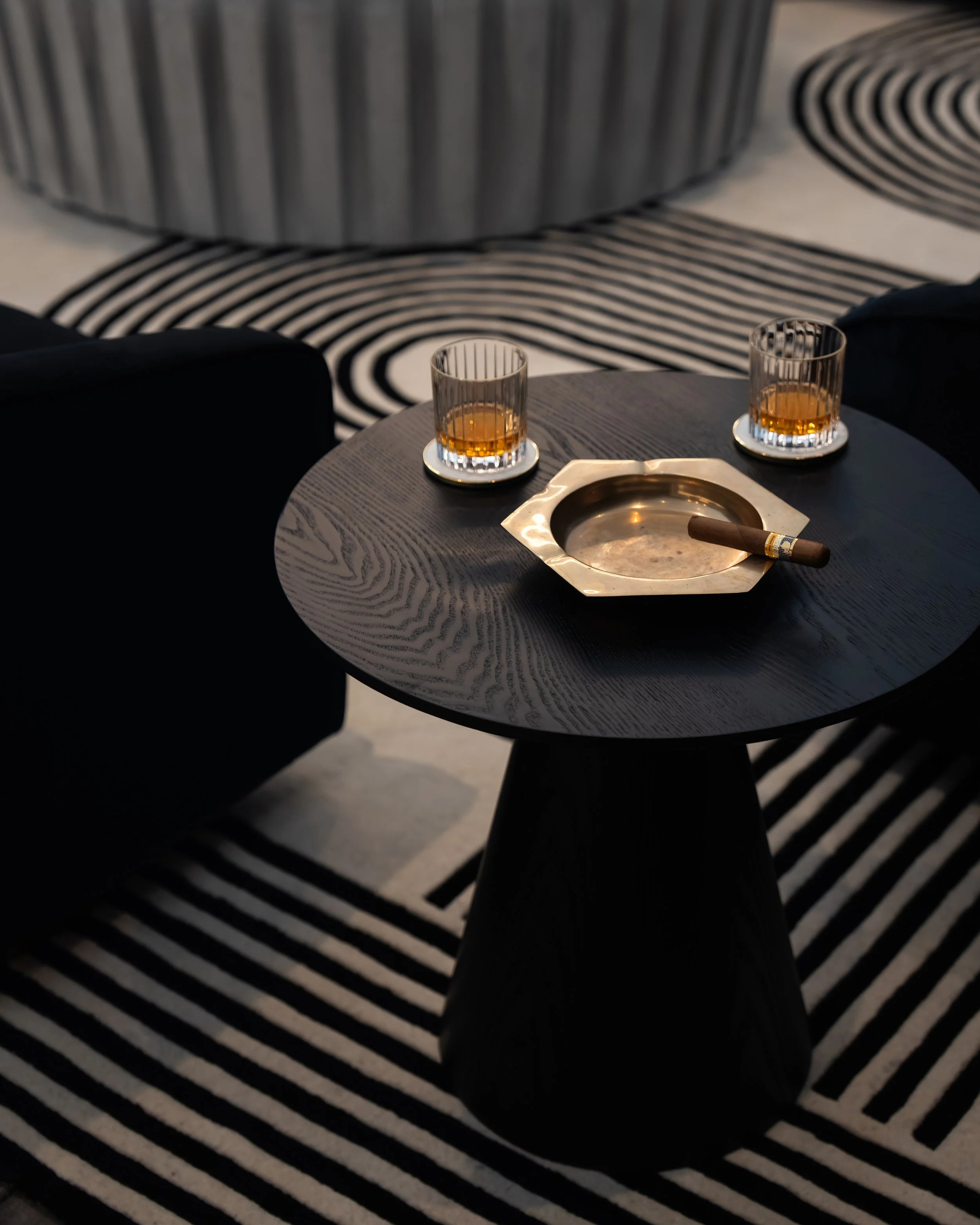 A black round side table with two glasses of amber-colored drinks, a cigar on a metallic ashtray, set on a black and white striped rug in a contemporary interior.