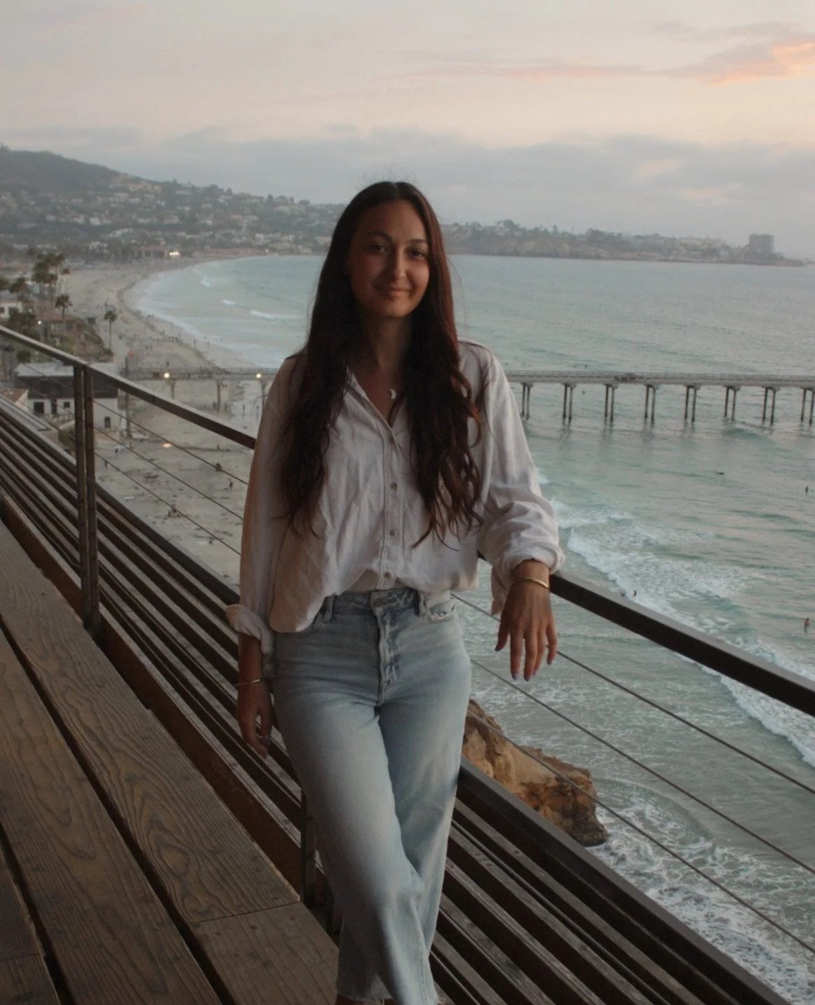 A woman in a white shirt and light blue jeans standing on a wooden deck overlooking the ocean during sunset, with a pier extending into the water and a hilly coastline in the background.