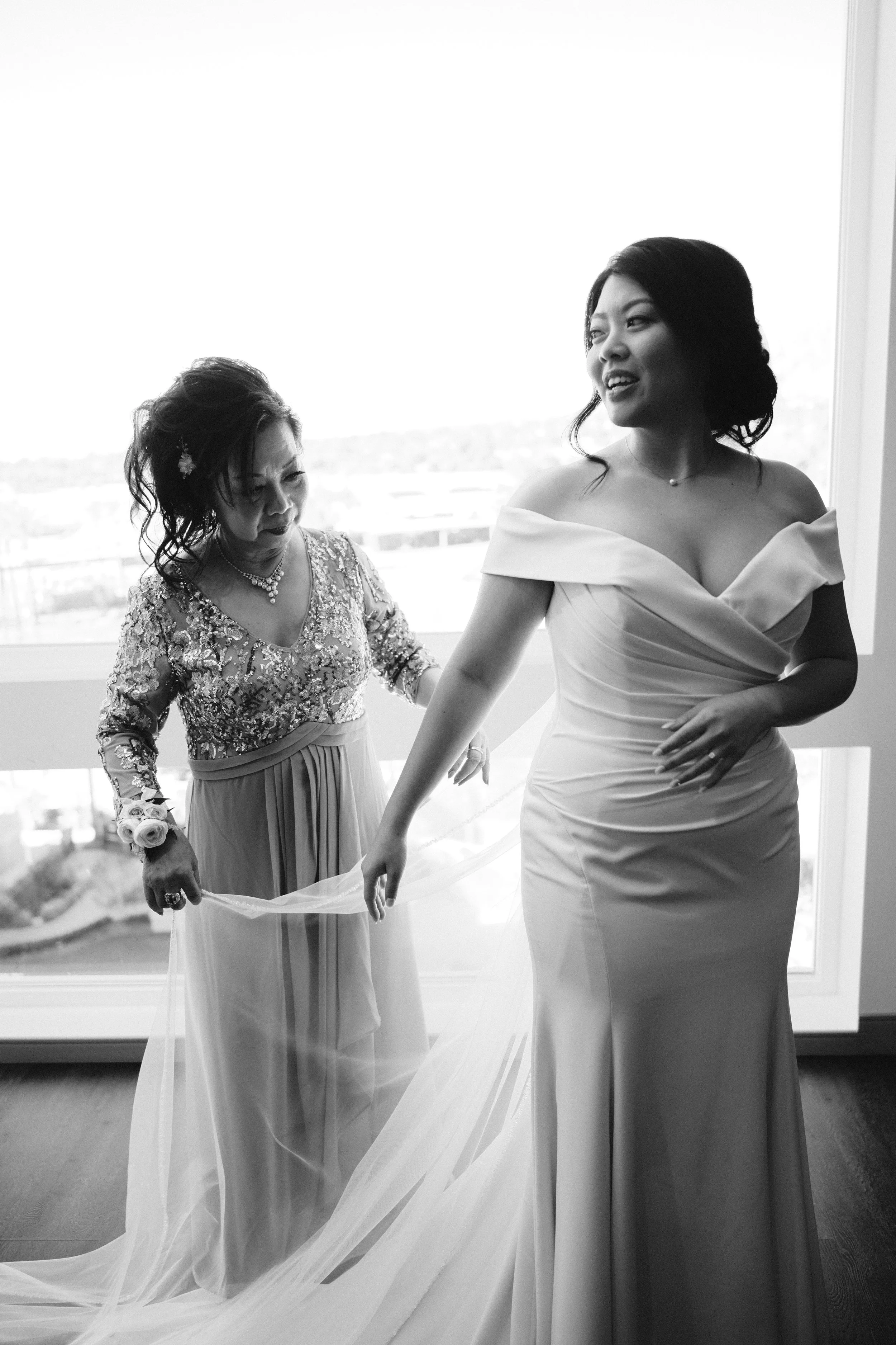 A bride in a wedding dress is assisted by another woman in an elegant gown, likely her mother, during a wedding preparation in a room with large windows.