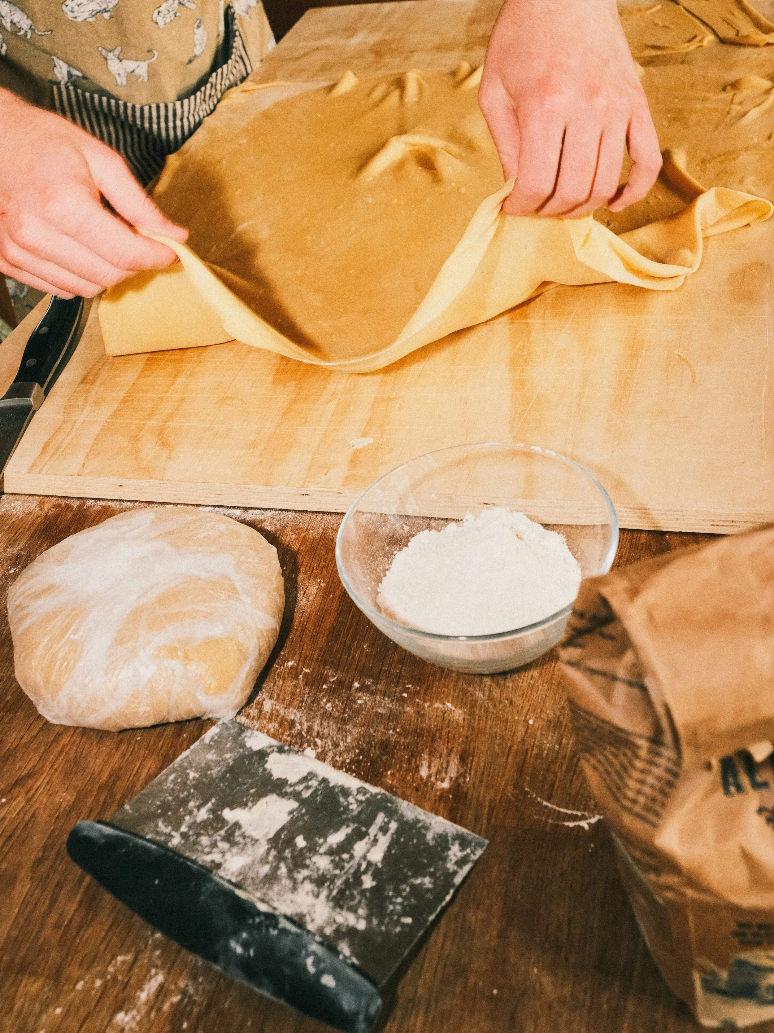 Person folding a sheet of rolled-out pie dough on a wooden kitchen counter with a bowl of flour, a ball of dough, a dough scraper, and a bag of flour nearby.