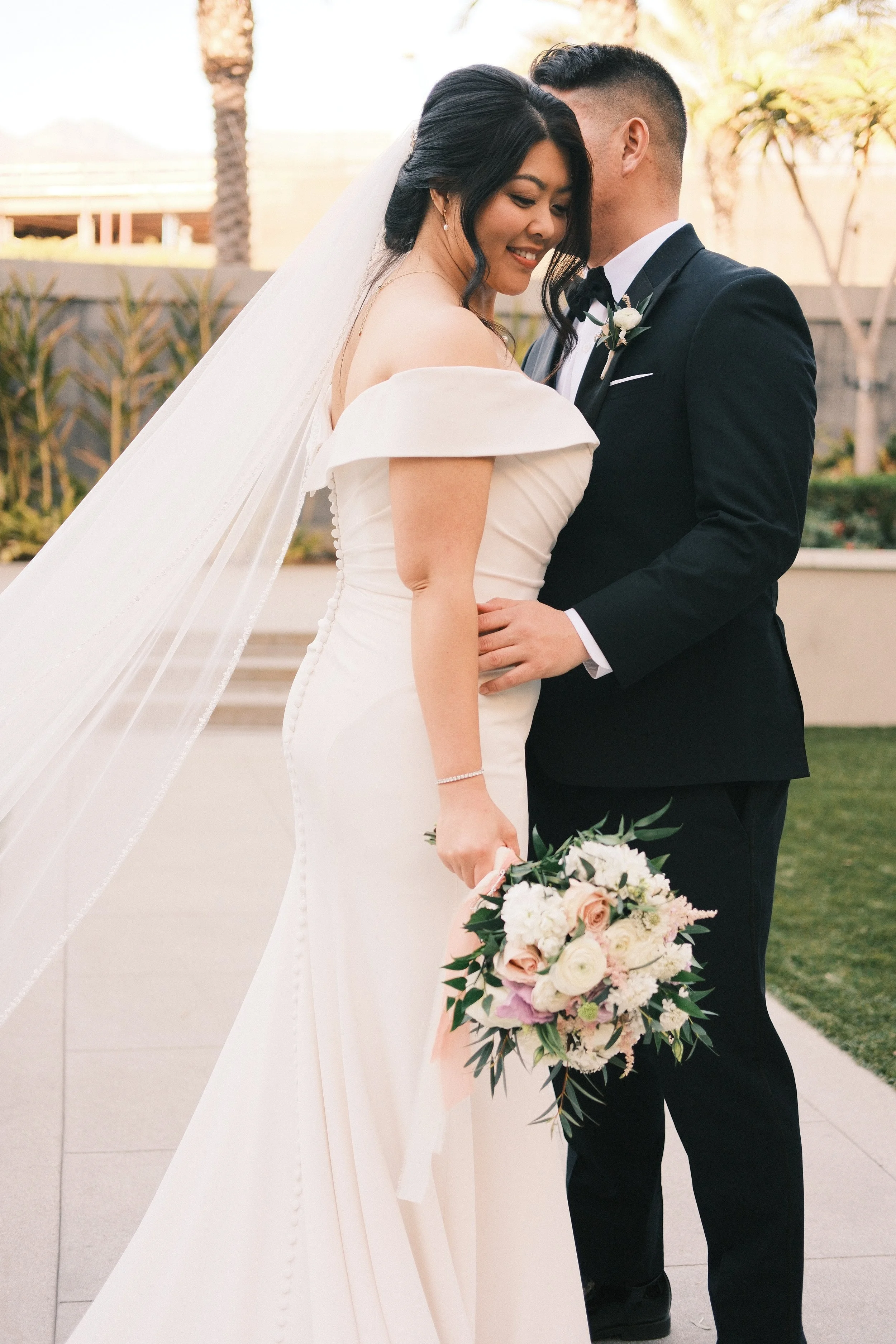 Bride in a white off-shoulder wedding gown holding a bouquet standing close with groom in a black tuxedo, outdoors with trees and buildings in the background.