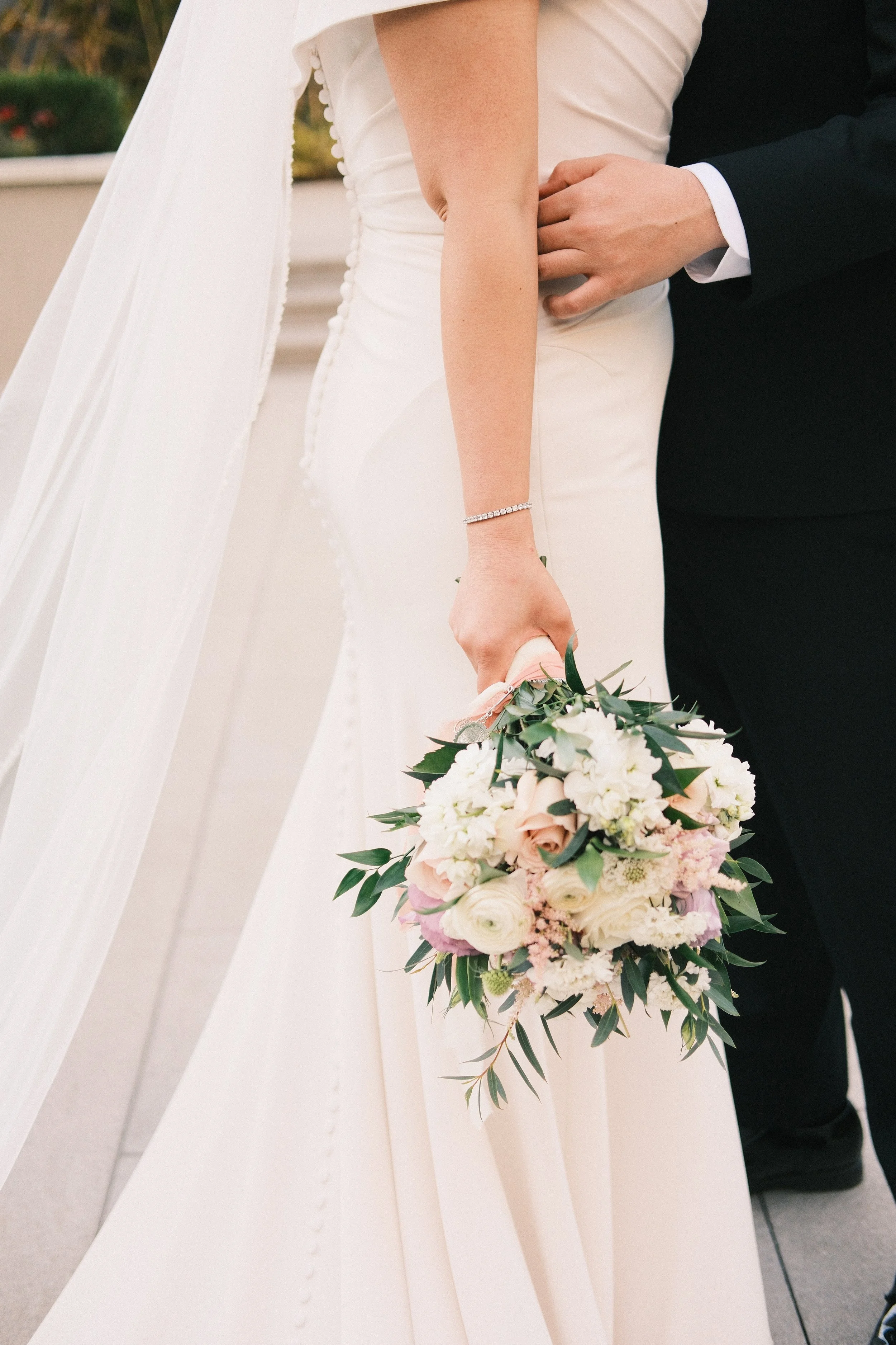 A bride holding a bouquet of white and light pink flowers, with a groom standing close by. The bride is wearing a white wedding dress with buttons down the back and a long veil.