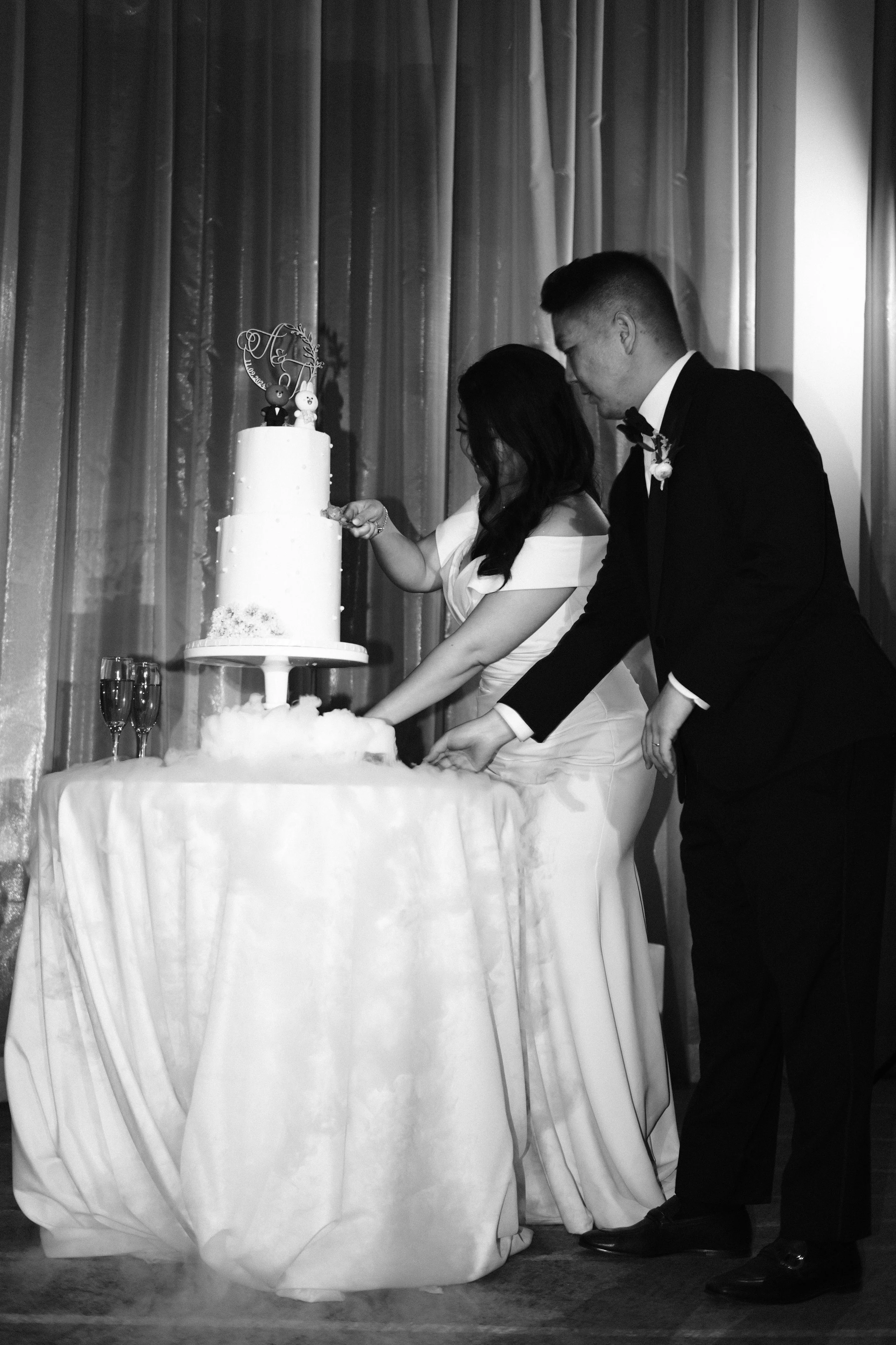 A bride and groom cut a wedding cake at their reception. The cake is multiple tiers with decorative elements, and the couple is dressed in formal wedding attire, standing in front of a curtain backdrop.