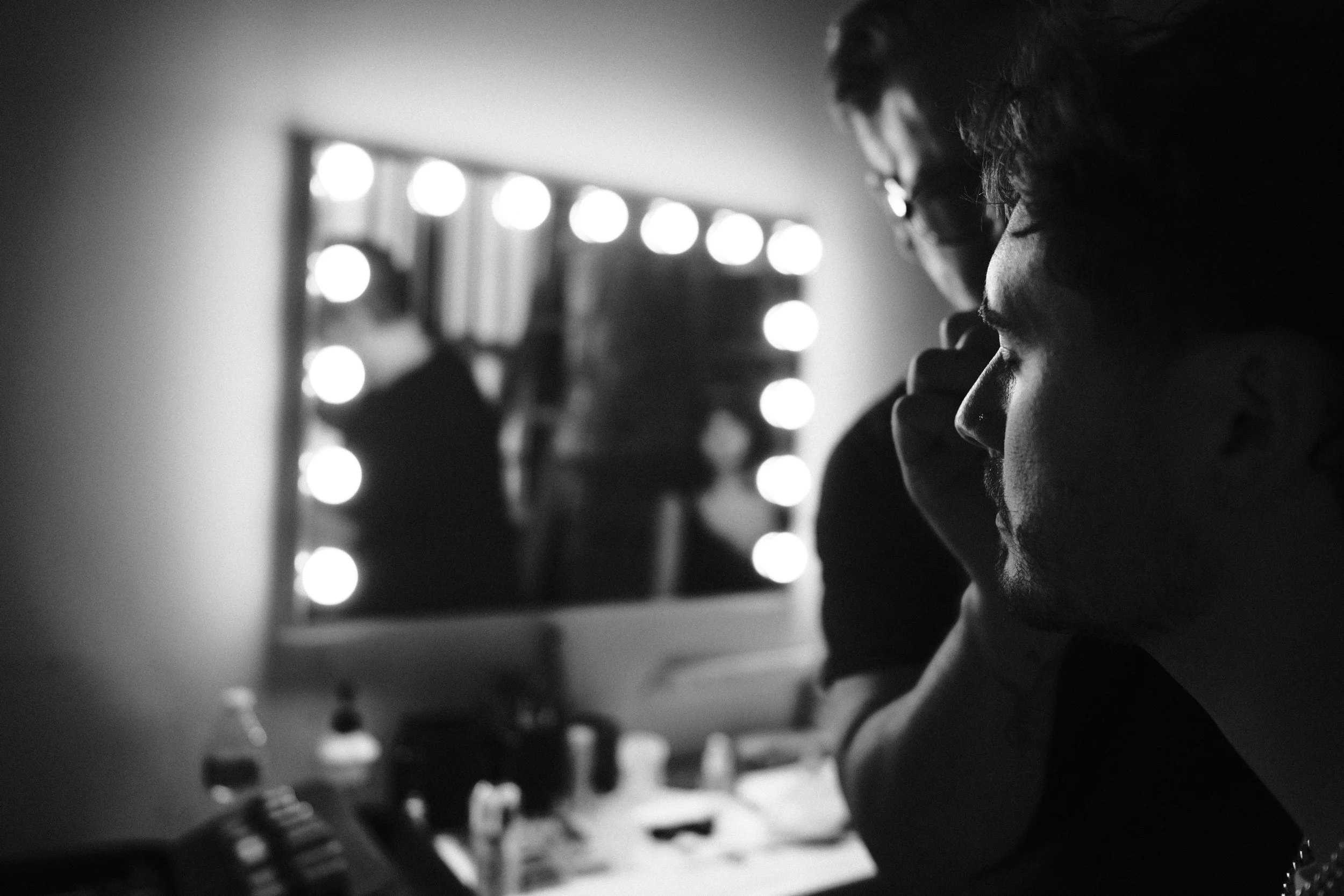 Two men with glasses looking into a mirror with lights around it, in a black-and-white photo.