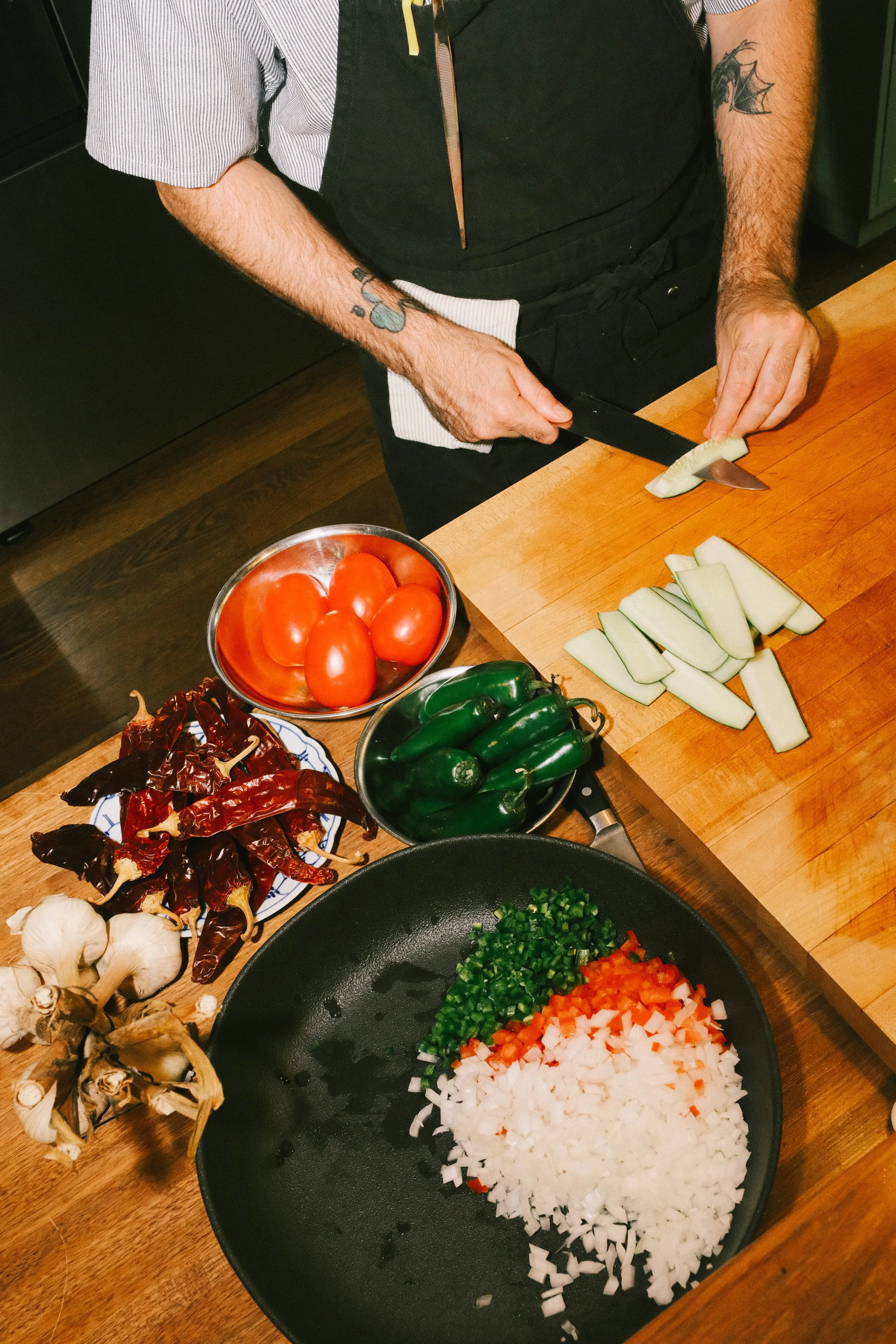 Person chopping a melon on a wooden cutting board surrounded by fresh vegetables including tomatoes, jalapeños, garlic, dried chili peppers, and diced onions and peppers in a black skillet.