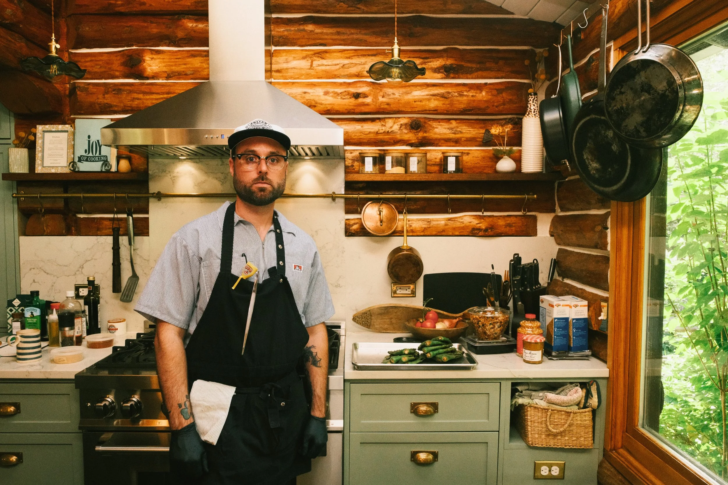 Man in apron and glasses standing in a rustic kitchen with wood walls, hanging pans, and various kitchen items, near a large window showing greenery outside.