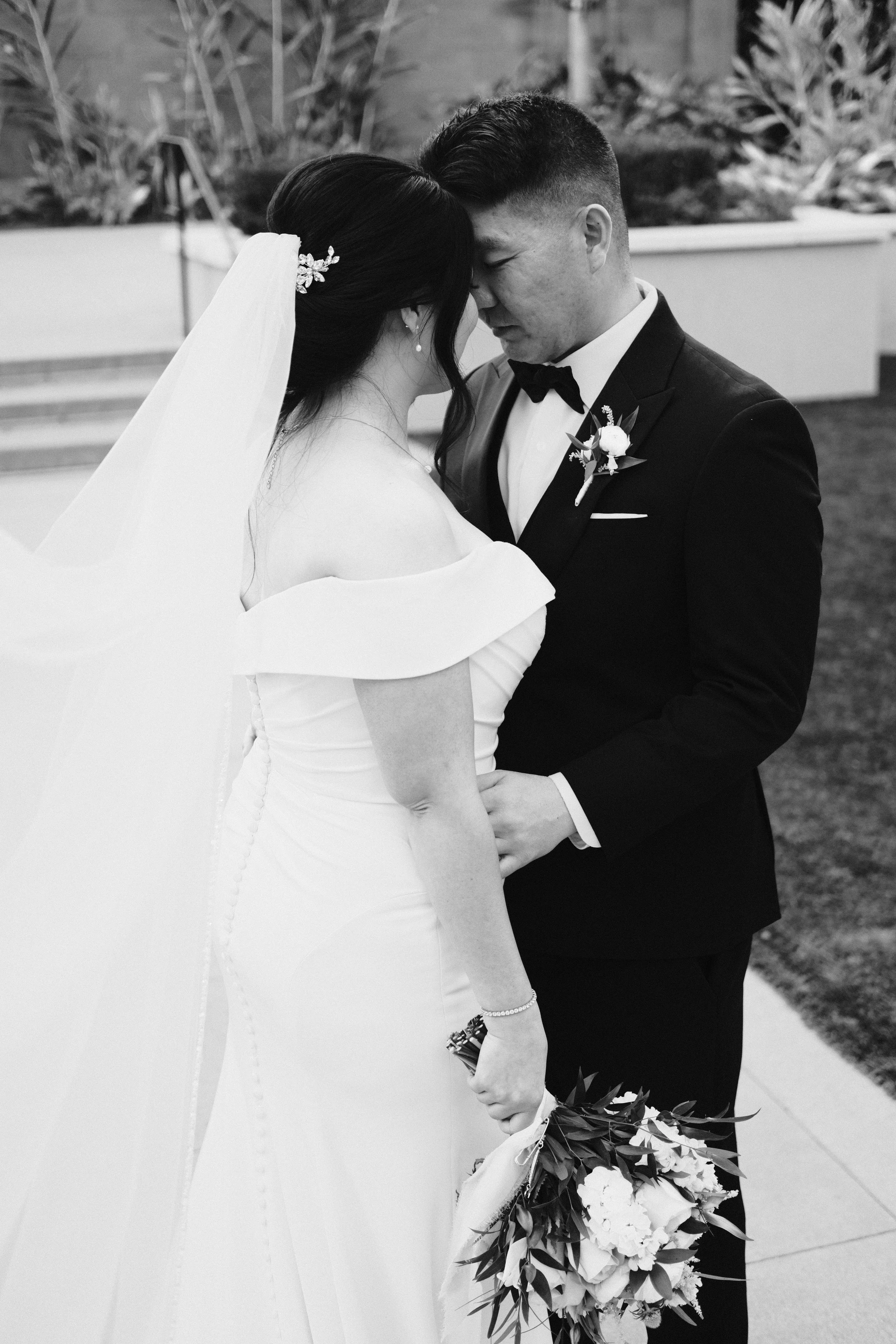 Black and white photo of a bride and groom holding hands and touching foreheads during their wedding.