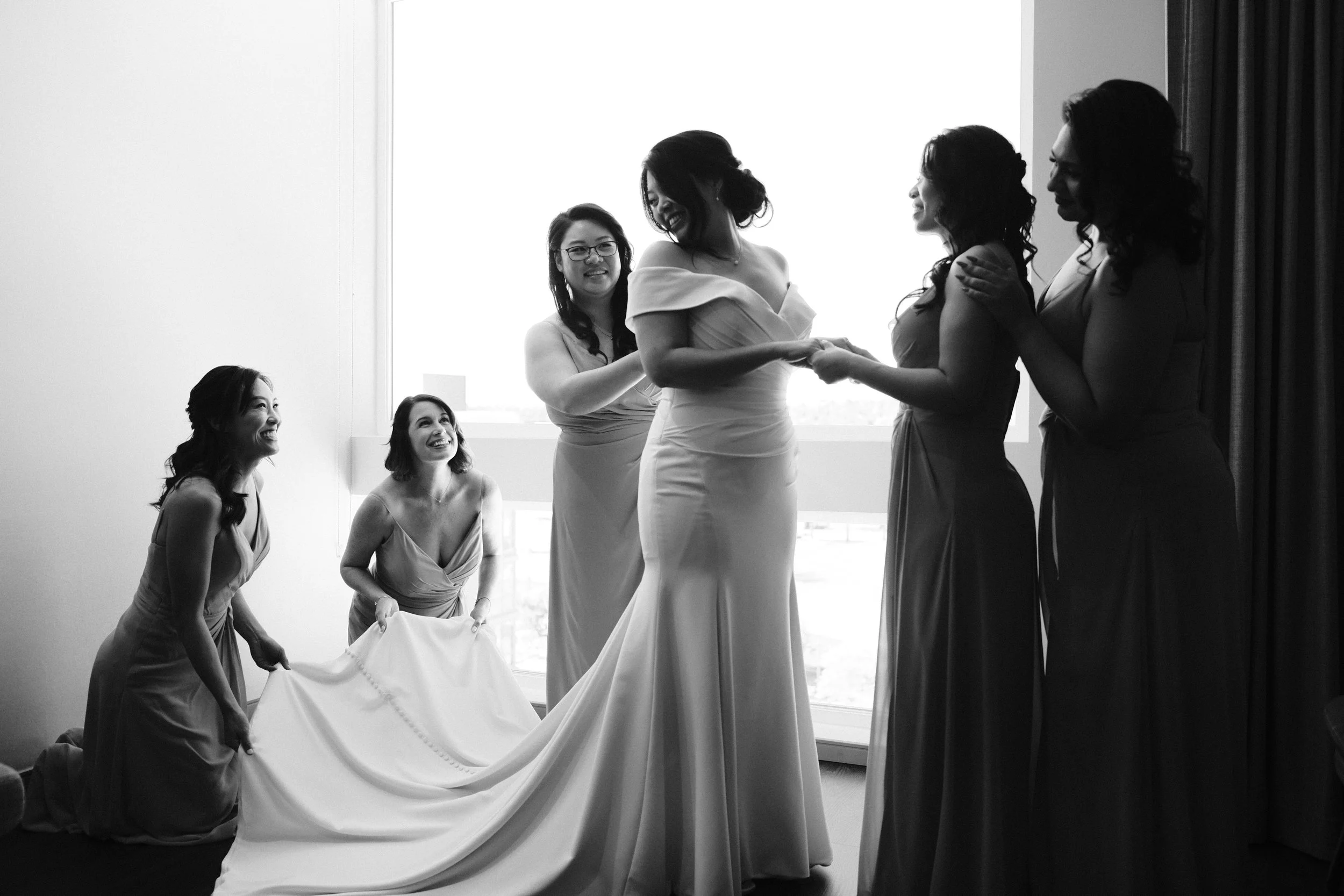 A bride and her bridesmaids in a black-and-white photo, with some standing and some kneeling, holding the hem of the bride's wedding dress in front of a large window.