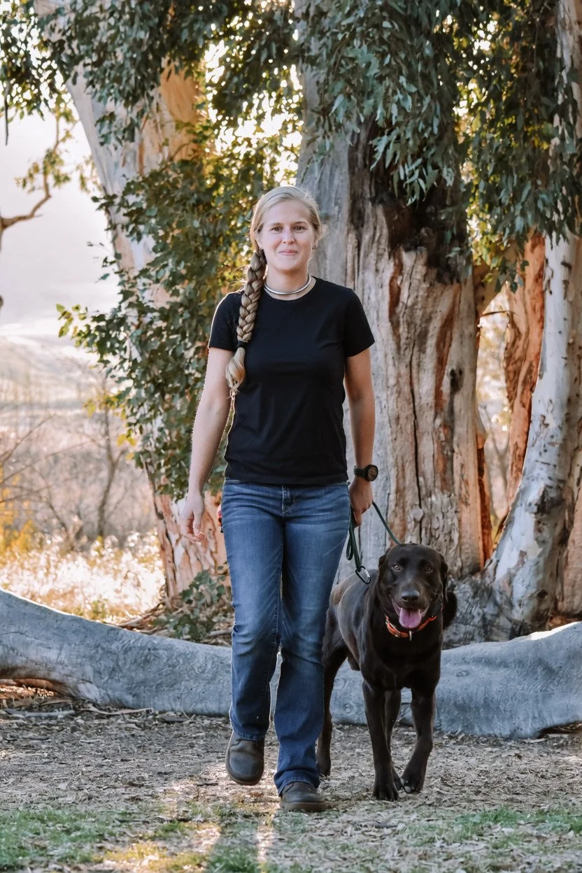 Katelyn standing with her chocolate Labrador, Grizzly, on a leash beneath trees.