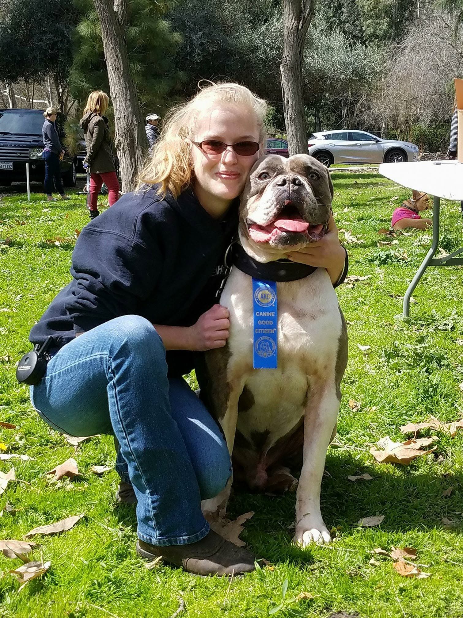 Katelyn posing outdoors with her American Bulldog, Moose, wearing an AKC Canine Good Citizen ribbon.