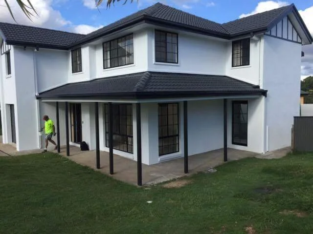 Two-story house with a black roof, white walls, and multiple windows. A covered patio with black support posts extends around part of the house. A person in a neon green shirt walks along the patio.