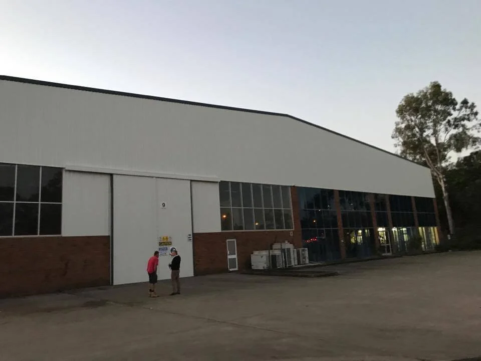A large industrial or warehouse building with a white roof and brick walls, two people standing near the entrance, and a few trees in the background at dusk.