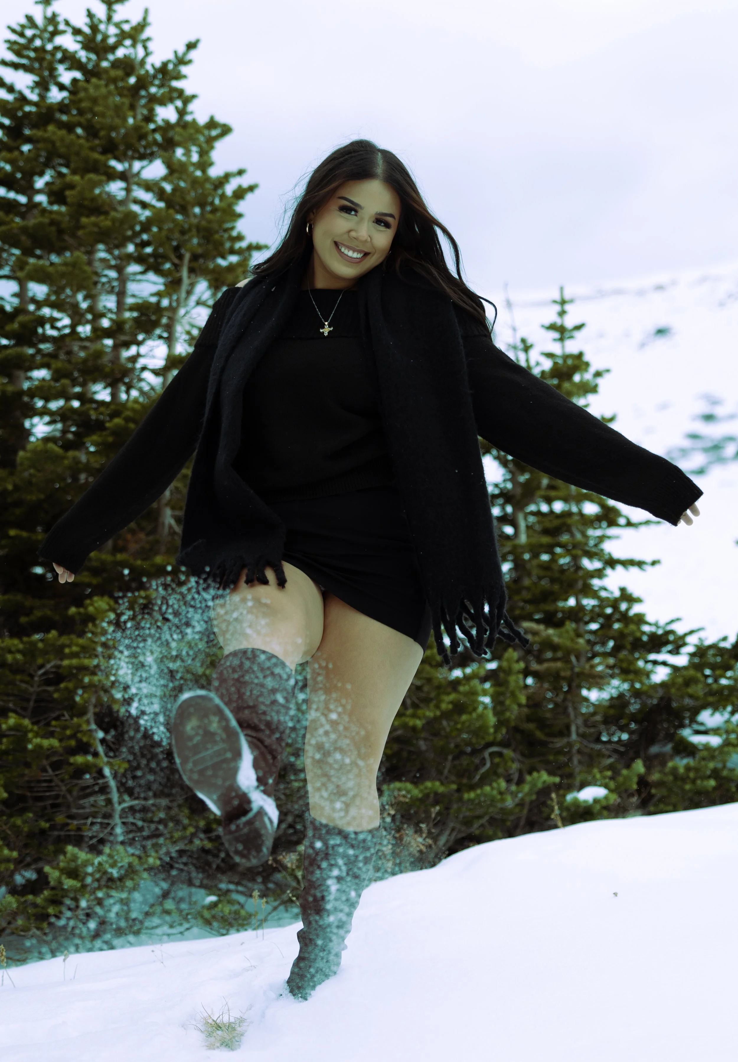 A woman in a black outfit and boots happily playing in the snow in a forested area with pine trees.