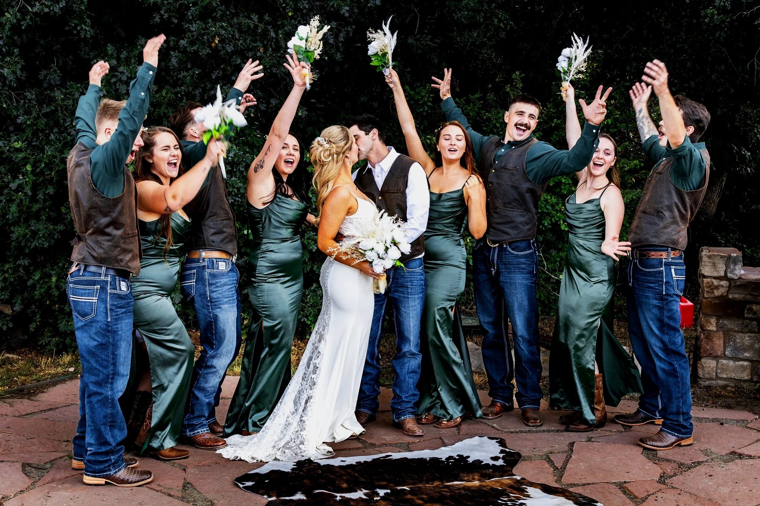 A group of wedding friends celebrating outdoors, with the bride and groom kissing in the center. The friends are raising their hands, cheering, and holding bouquets. They are dressed in coordinated outfits with the women in green dresses and the men 