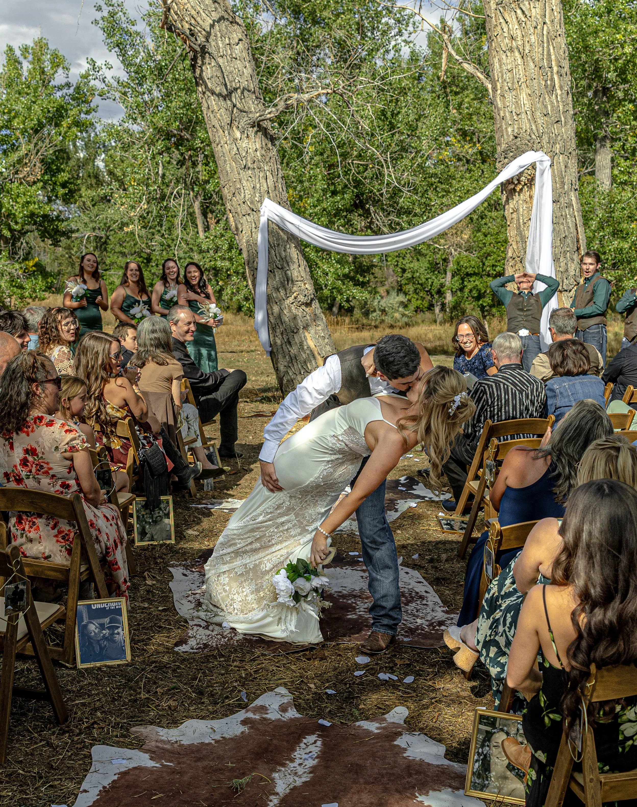 A couple kisses during their outdoor wedding ceremony surrounded by guests, trees, and a white draped fabric arch.