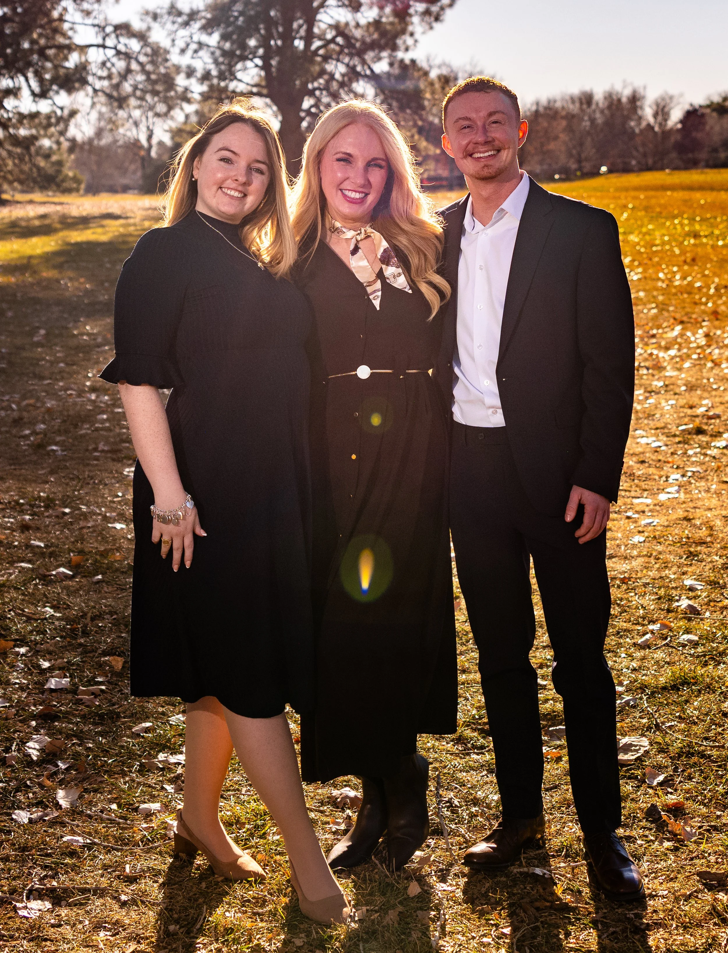 Three people standing outdoors in sunlight, smiling at the camera. They are dressed semi-formally with a setting of trees and grass in the background.