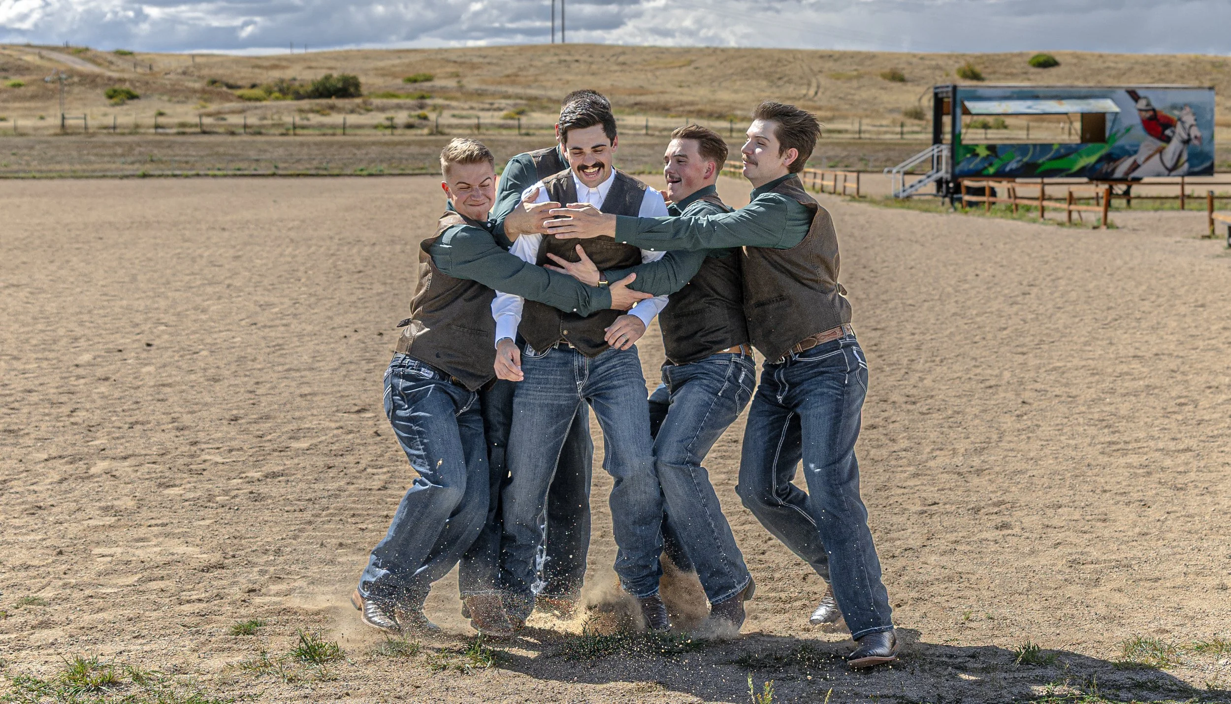 A group of five young men dressed in casual and western-style clothing are joyfully hugging and lifting each other in a dirt field.