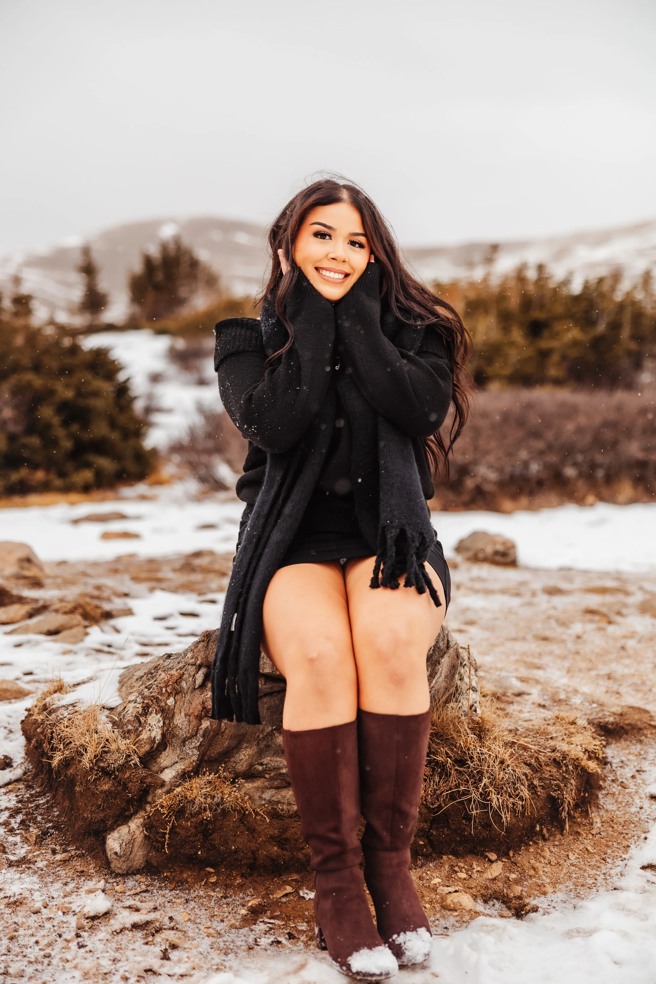 A woman with long dark hair smiling and sitting on a rock in a snowy outdoor landscape, wearing a black sweater, scarf, shorts, and brown boots.