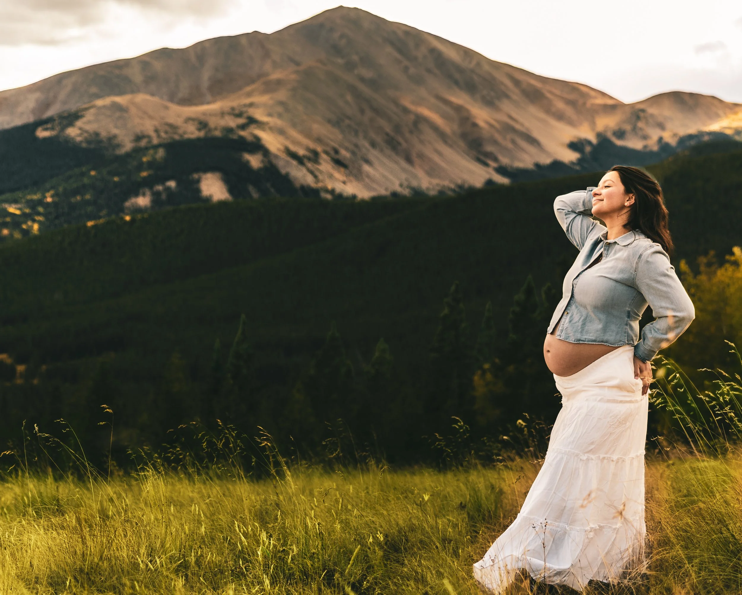A pregnant woman standing in a grassy field with mountains in the background, wearing a denim jacket and a white flowing skirt, with her eyes closed and one hand on her head.