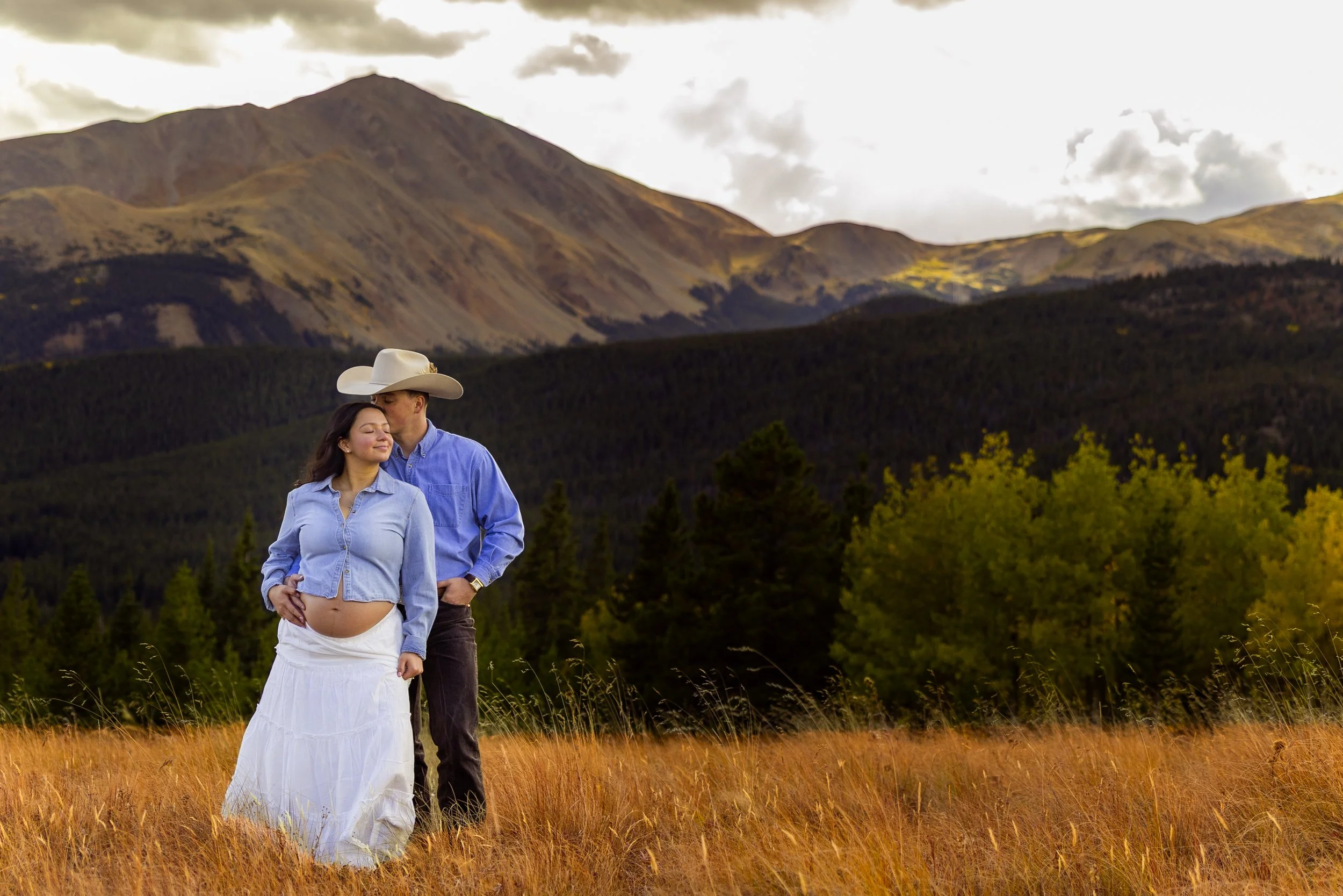A pregnant woman and a man standing together in a field with mountains in the background. The woman is smiling with her eyes closed, wearing a light blue denim shirt and a white skirt, while the man, dressed in a blue shirt, black jeans, and a cowboy