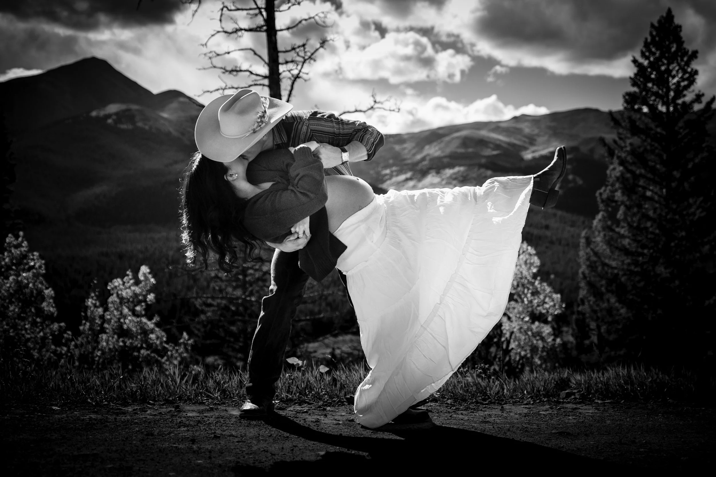 A black and white photo of a man holding a woman in a dance pose outdoors with mountains, trees, and a cloudy sky in the background.